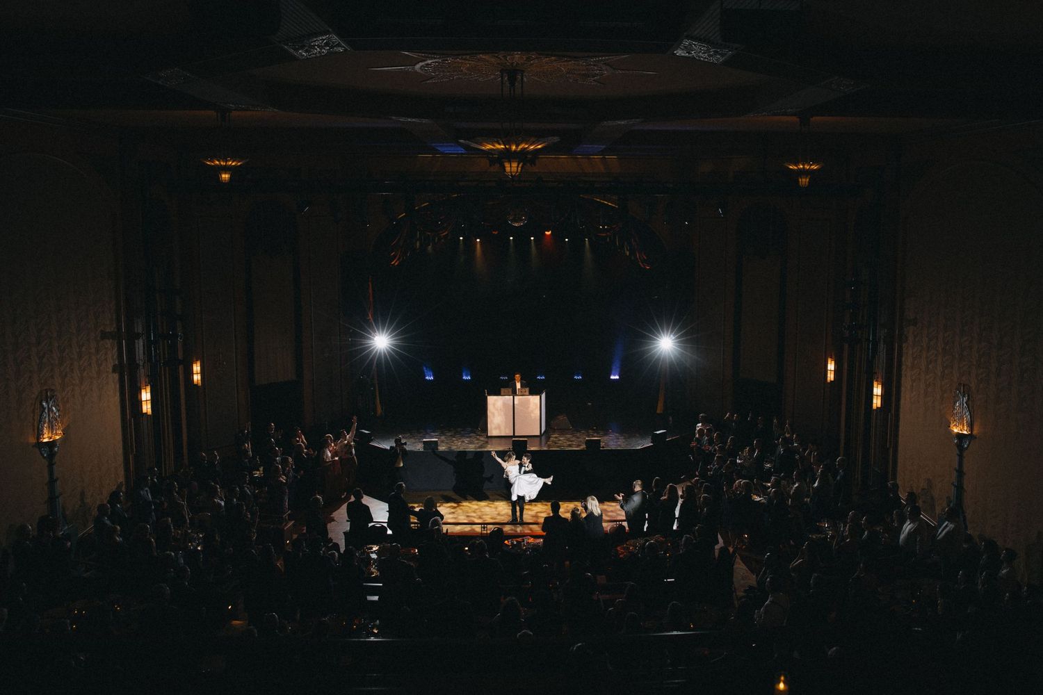 Wedding ceremony in a grand theater venue with guests seated and bright stage lights illuminating the couple.