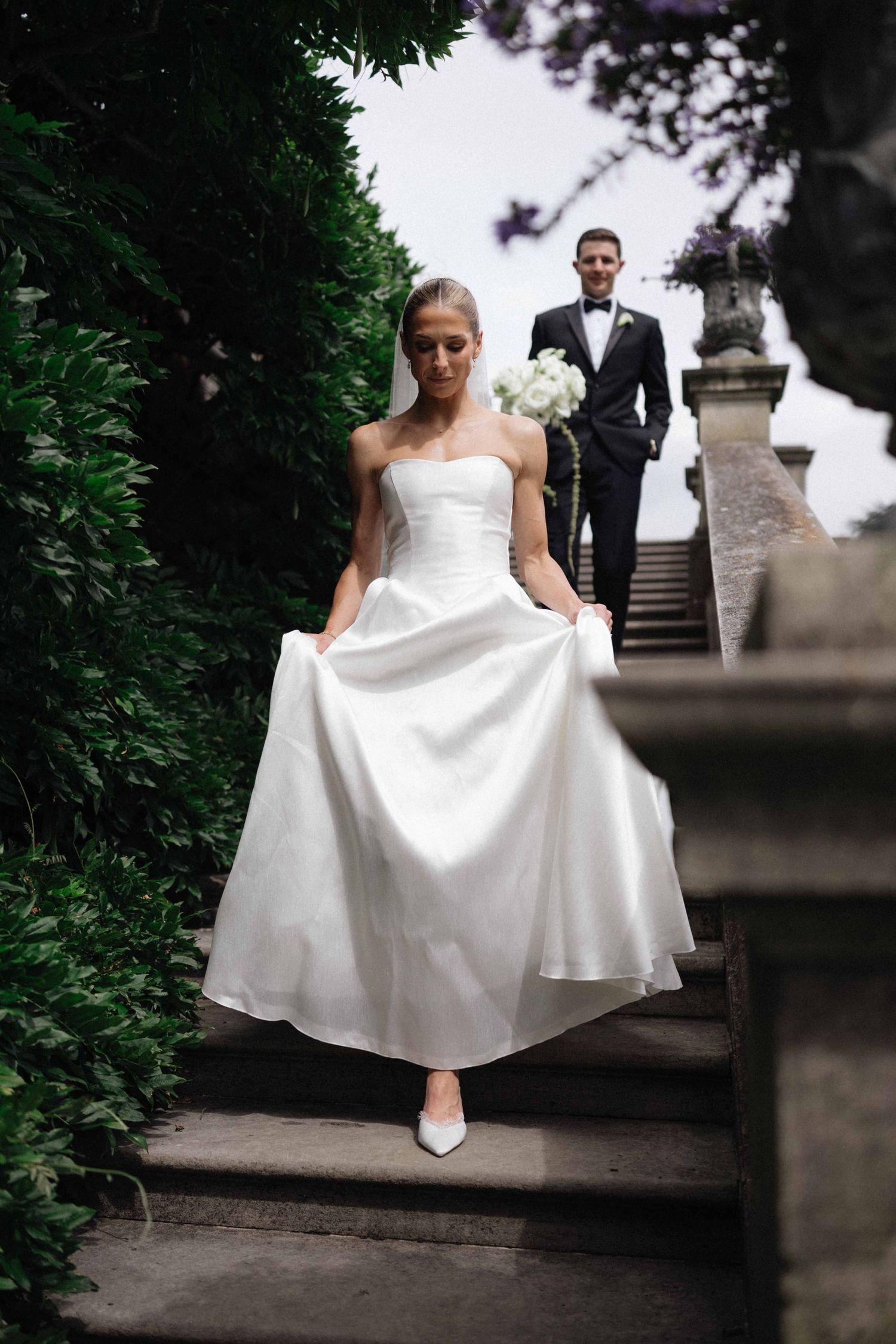 Bride in white strapless gown descends stone steps holding bouquet, groom in black tuxedo follows behind.