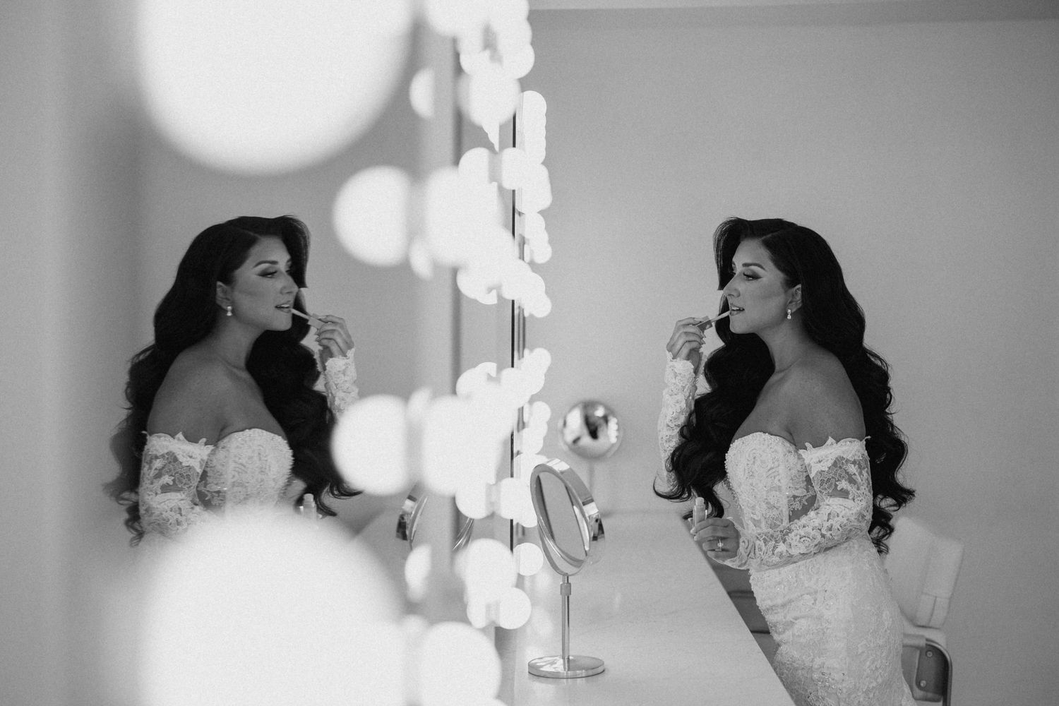 Black and white photo of a bride in lace gown applying lipstick before a Hollywood vanity mirror.