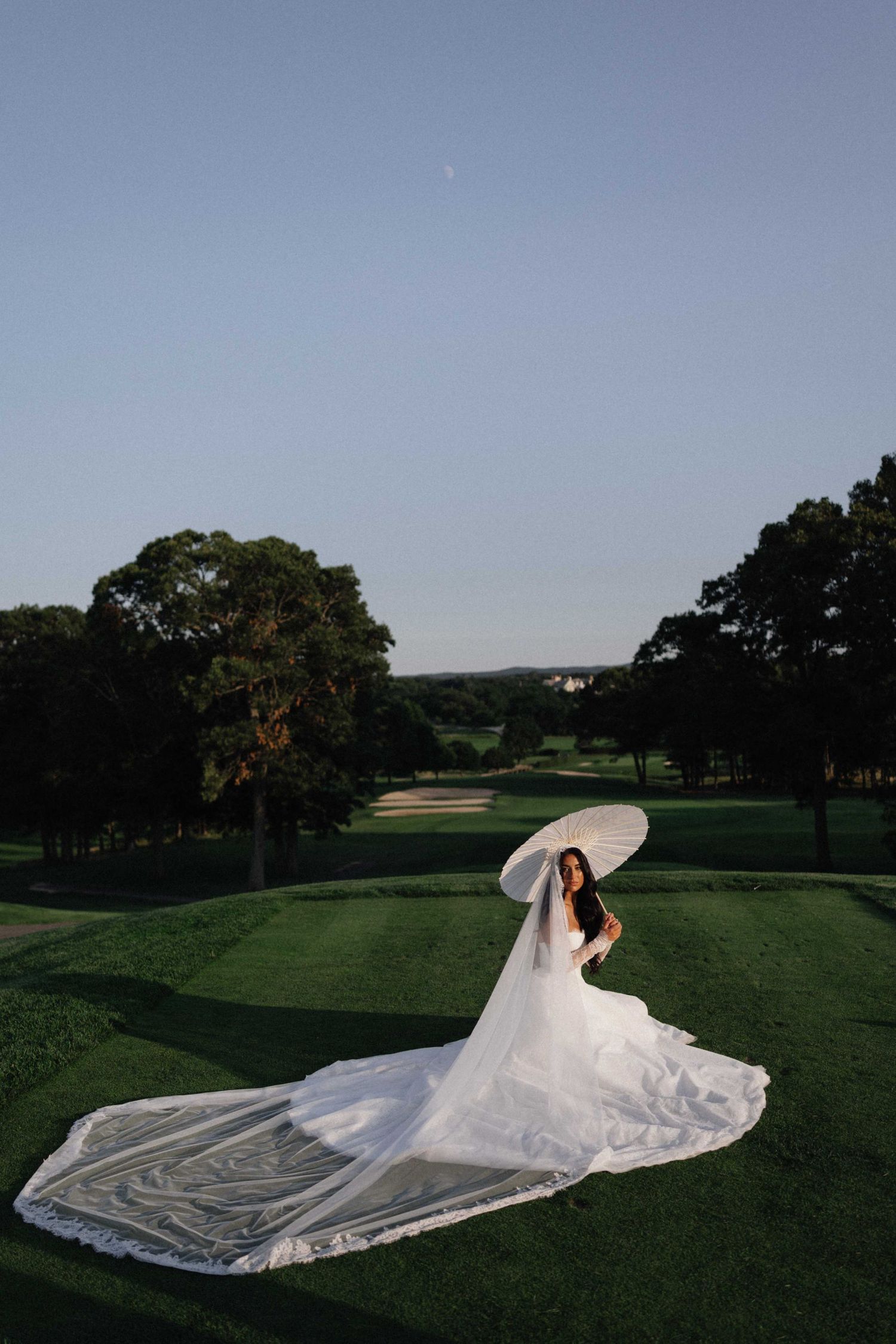 Bride in a flowing white gown with long train holds a parasol on a lush green golf course at dusk.