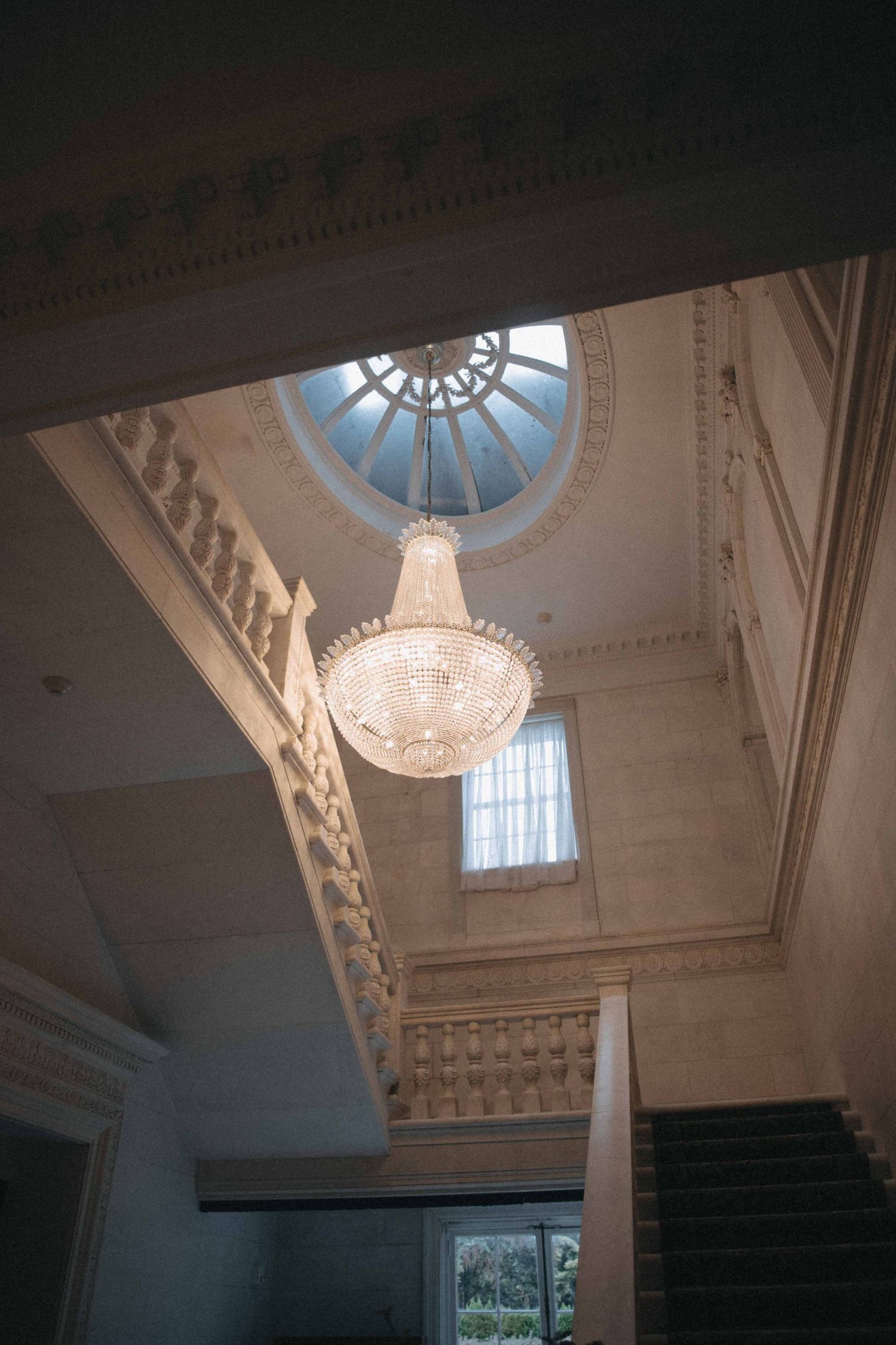Grand crystal chandelier hangs beneath ornate domed skylight in elegant historic venue with decorative moldings and staircase.