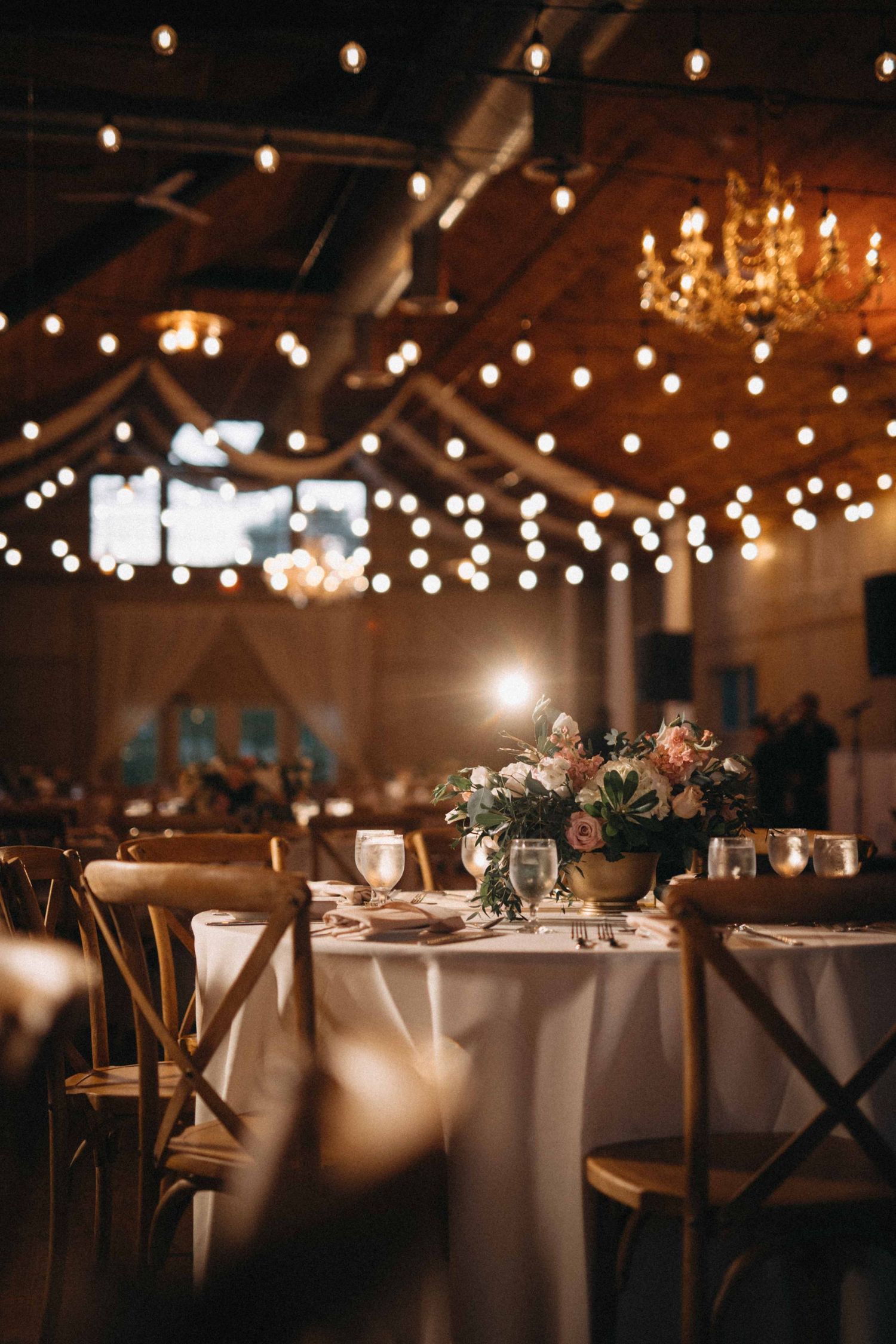 Elegant barn wedding reception with string lights, chandeliers, and floral centerpieces on white tables.