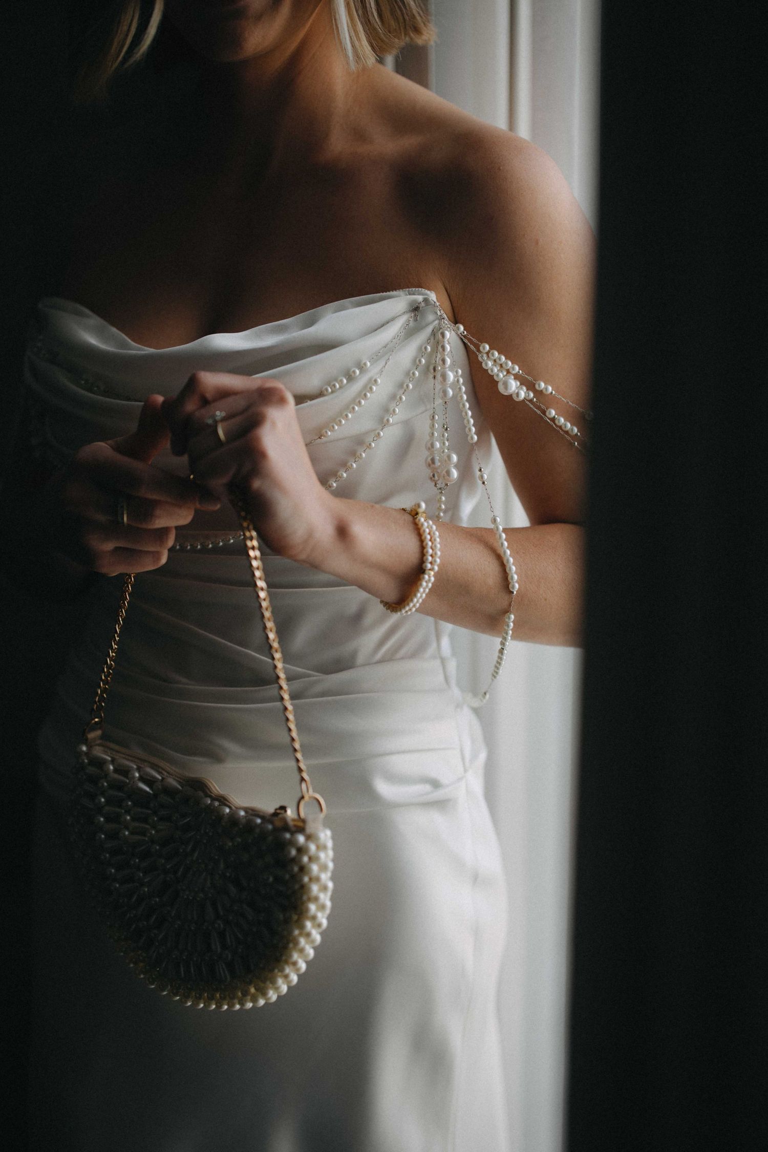 Bride in a strapless white gown holds a pearl-embellished clutch bag with delicate pearl strap details near a window.