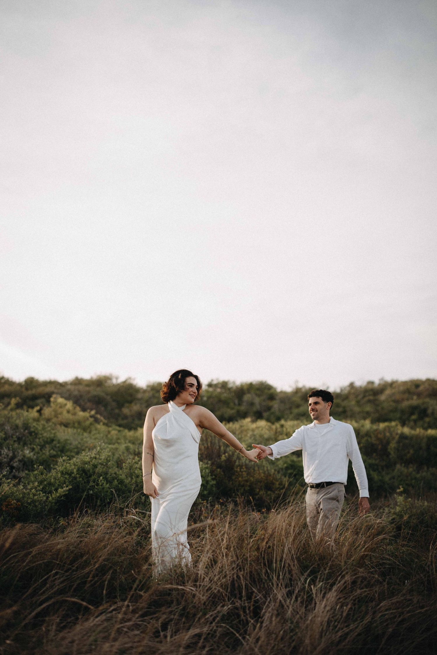 Couple holding hands walking through tall grass field, woman in white halter dress, man in white shirt and khakis.