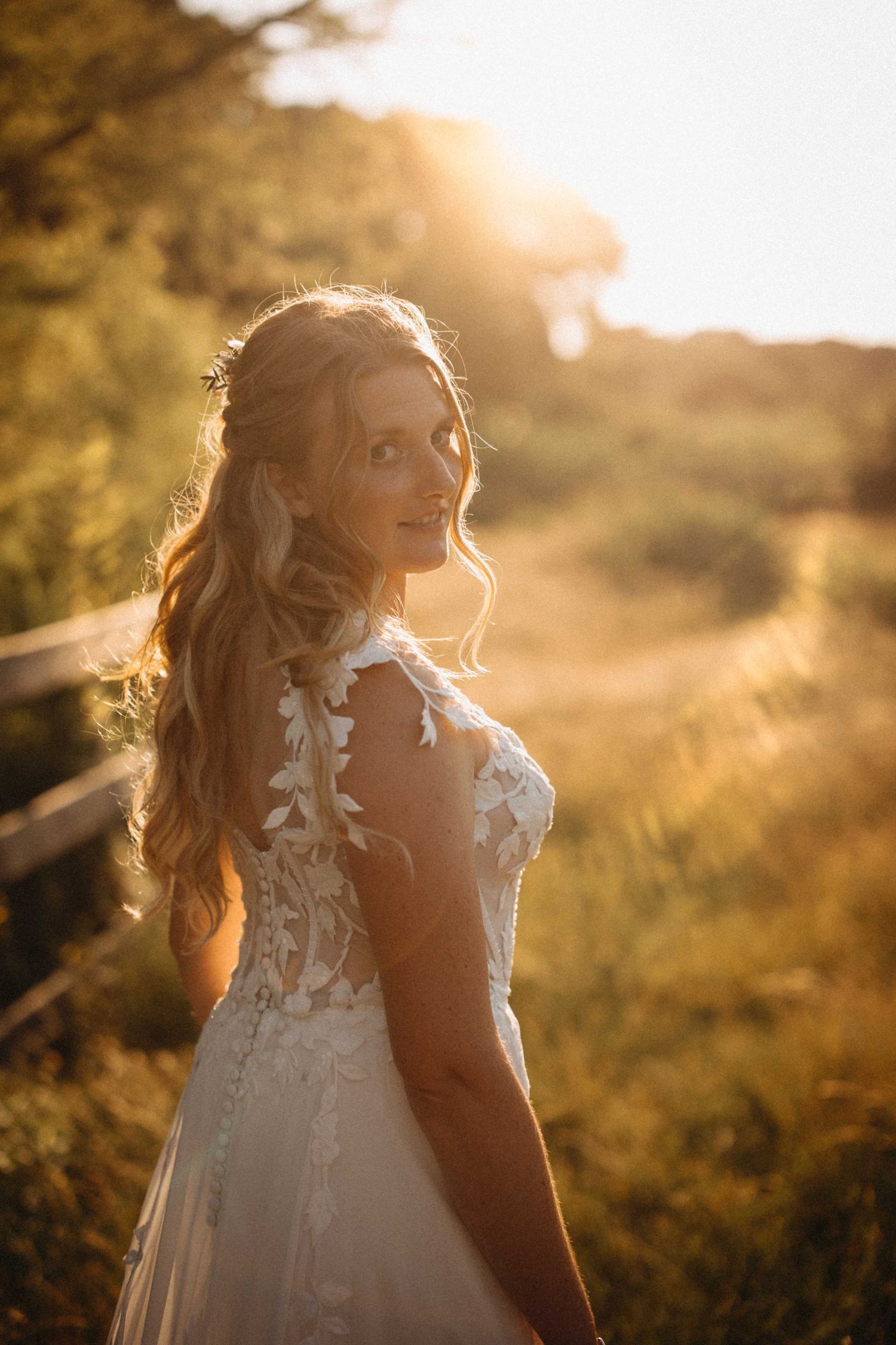 Blonde bride in a floral lace wedding dress glowing in warm golden sunset light outdoors.