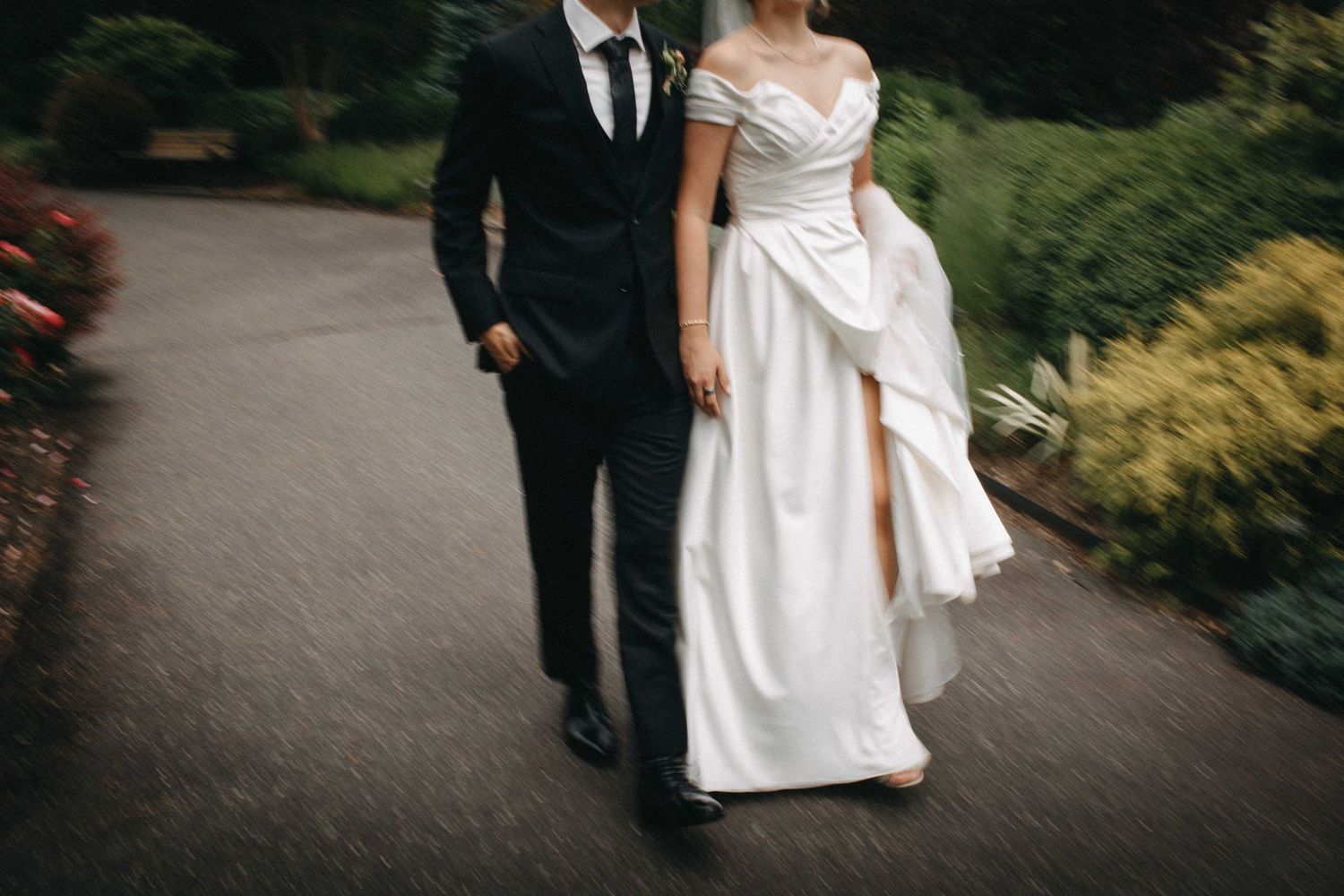 Bride in off-shoulder gown with slit and groom in dark suit walking on garden path.