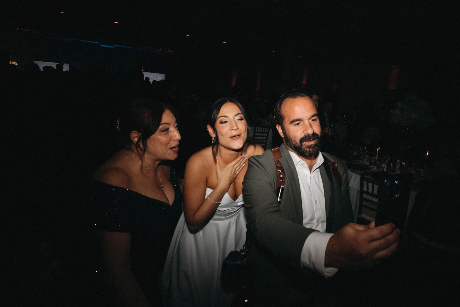 Bride in white dress posing for selfie with guests at wedding reception.
