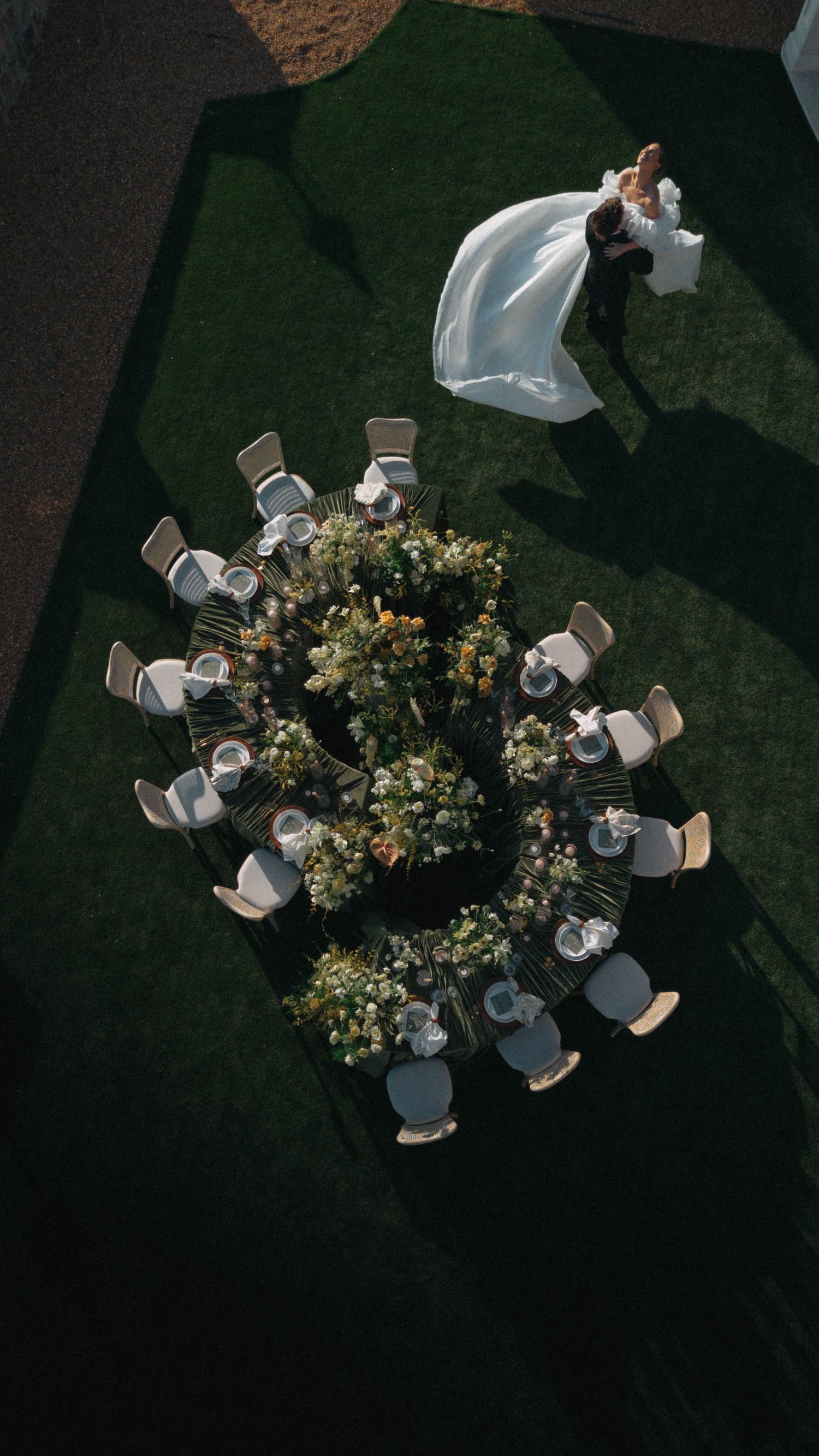 Aerial view of a circular floral wedding table arrangement on green grass with a bride and groom dancing nearby.