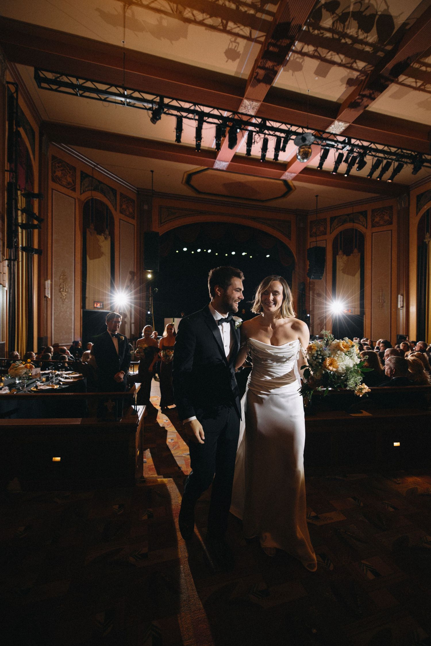 Bride and groom walk together in elegant ballroom wedding reception with ornate arched decor and guests dining.