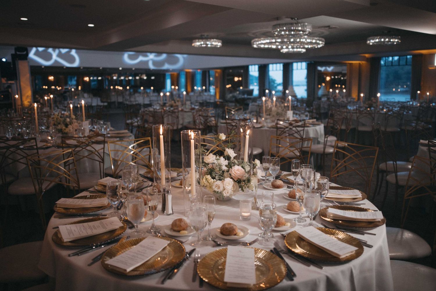 Elegant wedding reception hall with gold charger plates, candles, and crystal chandeliers overhead.