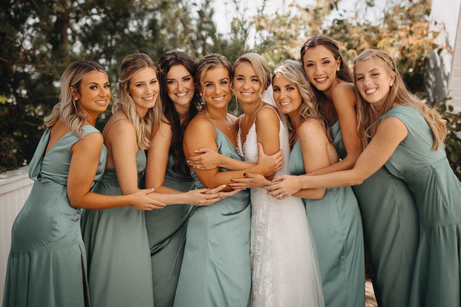 Bride surrounded by six smiling bridesmaids in sage green dresses in warm golden light.