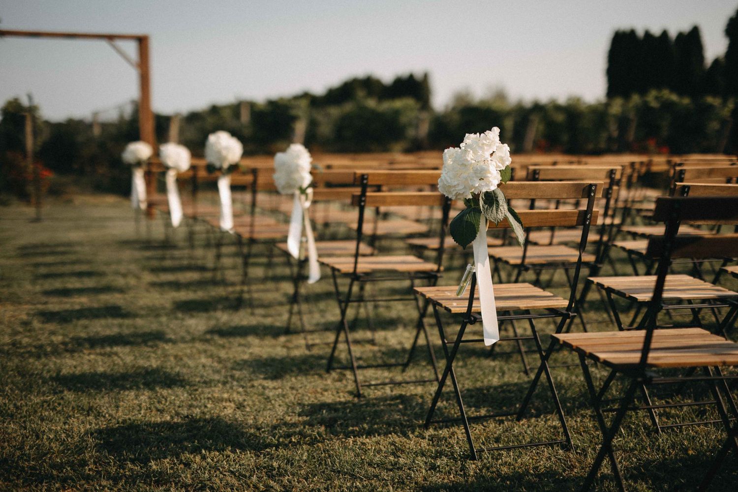 Elegant outdoor wedding ceremony chairs lined with white floral bouquets on a sunlit lawn.