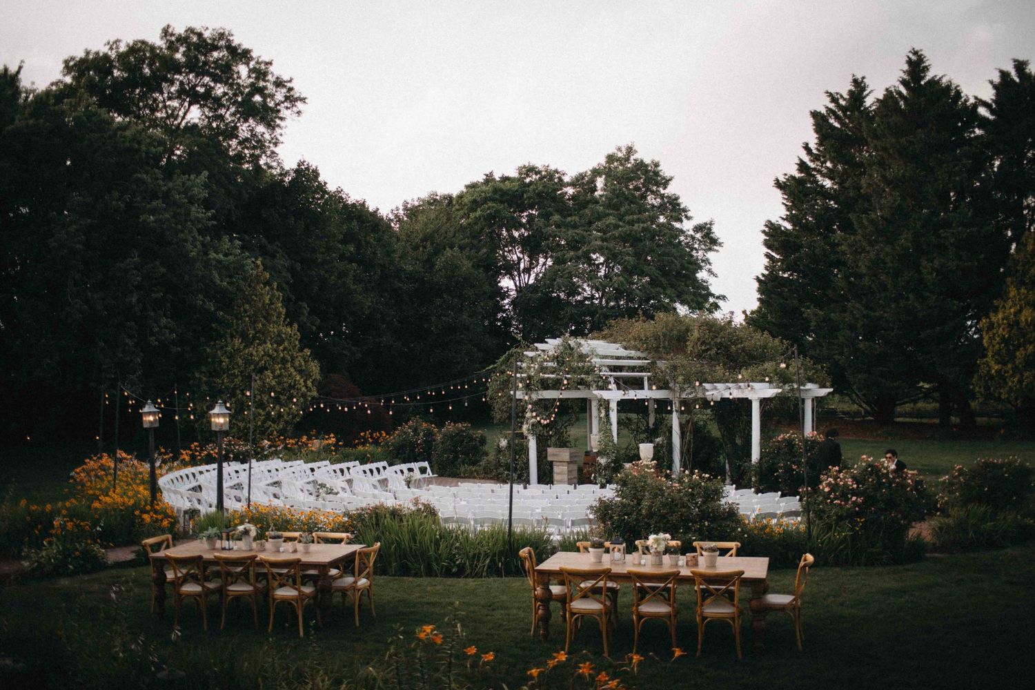 Outdoor wedding reception setup with wooden tables, string lights, and white pergola surrounded by lush greenery.