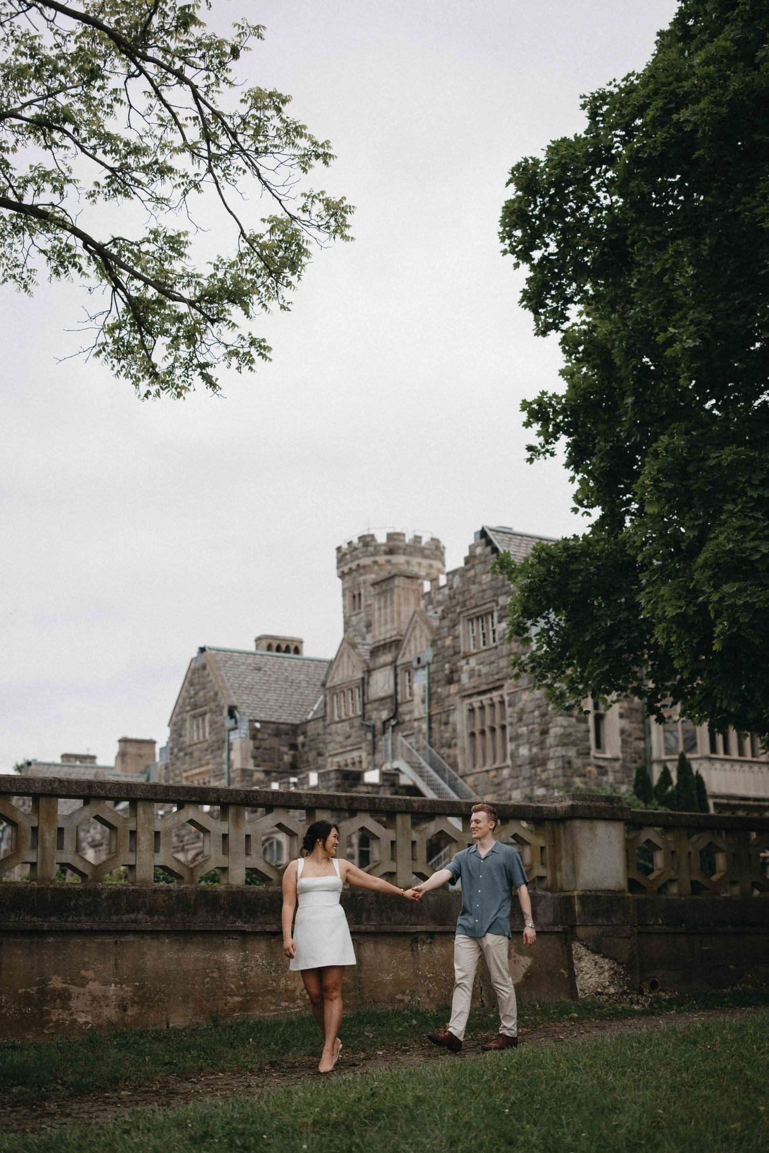 A couple holds hands in front of a grand historic stone castle surrounded by lush green trees.