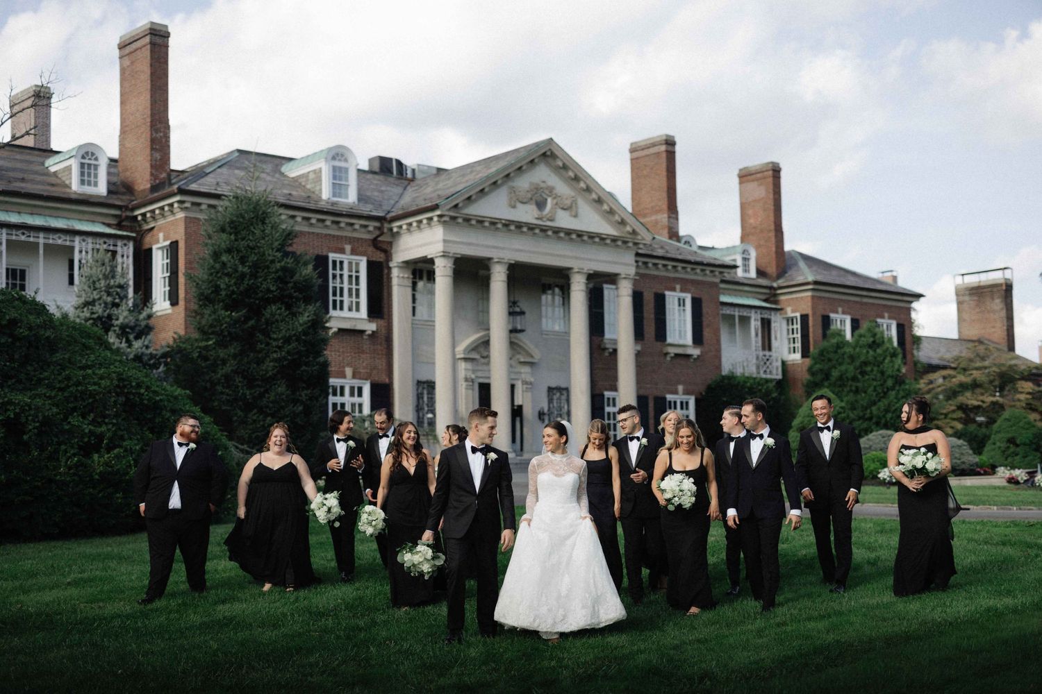 Wedding party in black attire poses with bride and groom on lawn of elegant brick colonial mansion.