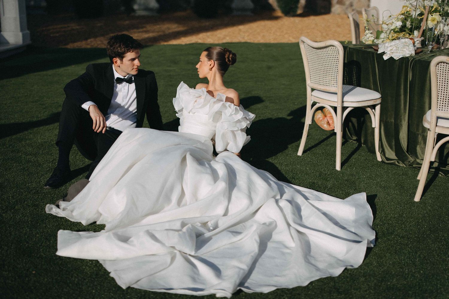 Bride in white ball gown and groom in tuxedo sitting together on green lawn at elegant outdoor wedding.