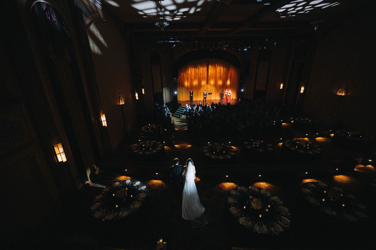 Bride walks down the aisle in a stunning dark wedding venue with glowing stage and candlelit round tables.