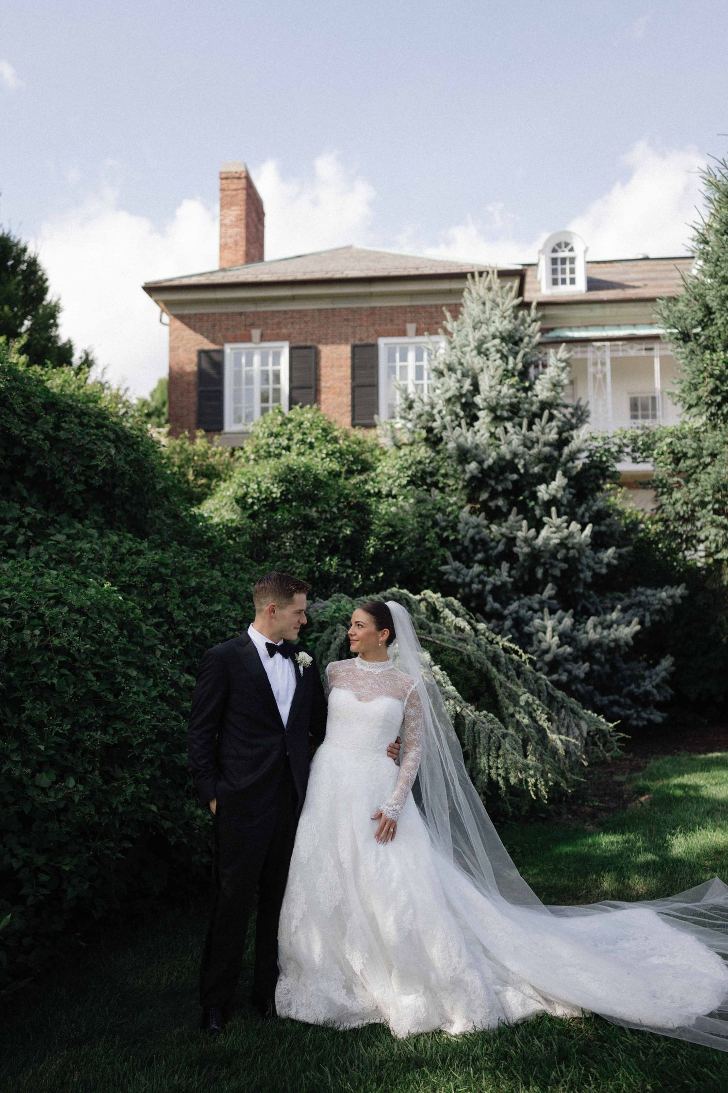 Bride and groom share a romantic moment in lush garden grounds before a classic brick estate on their wedding day.