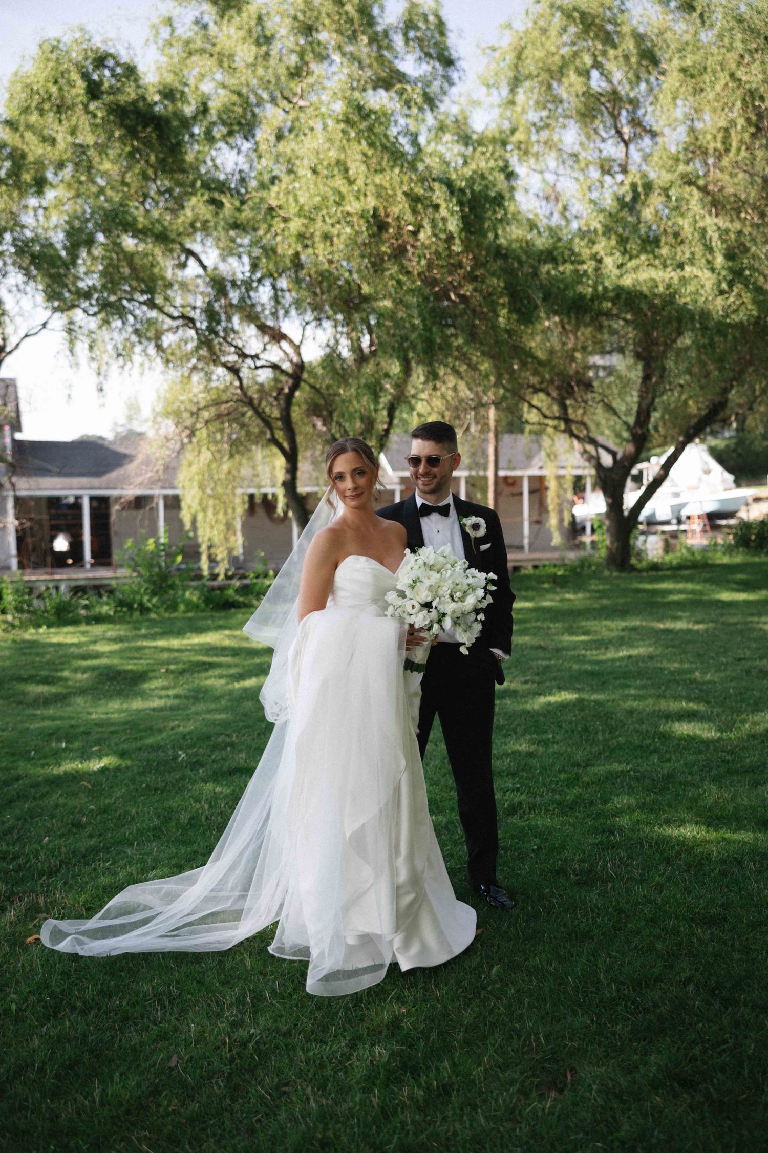 Bride in white strapless gown with veil and groom in black tuxedo posing on green lawn with trees behind them.