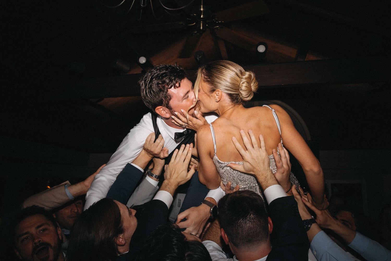 Bride and groom kissing while being lifted by wedding guests during energetic reception celebration.