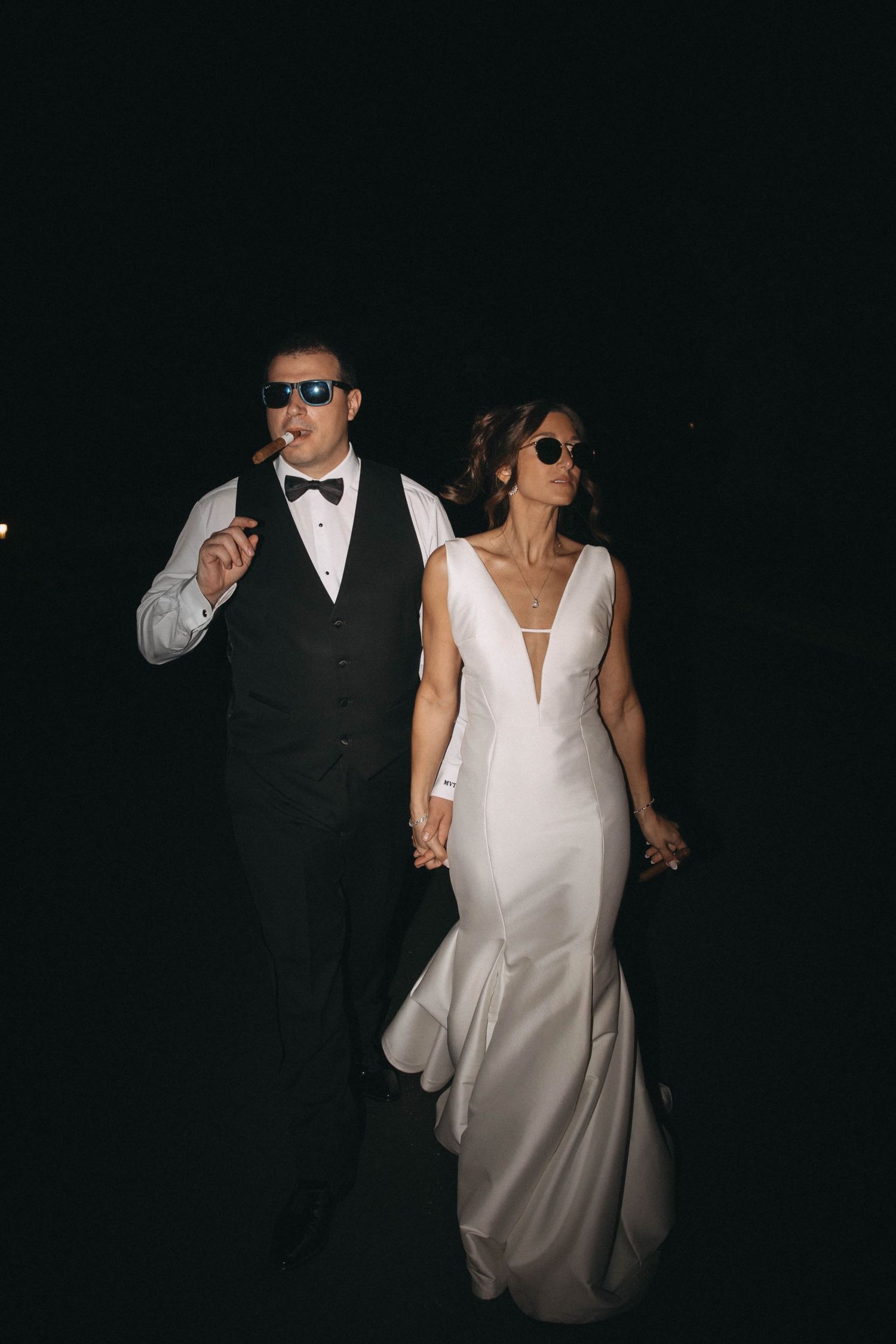 Bride and groom walking at night wearing sunglasses, groom smoking cigar in black tuxedo vest.