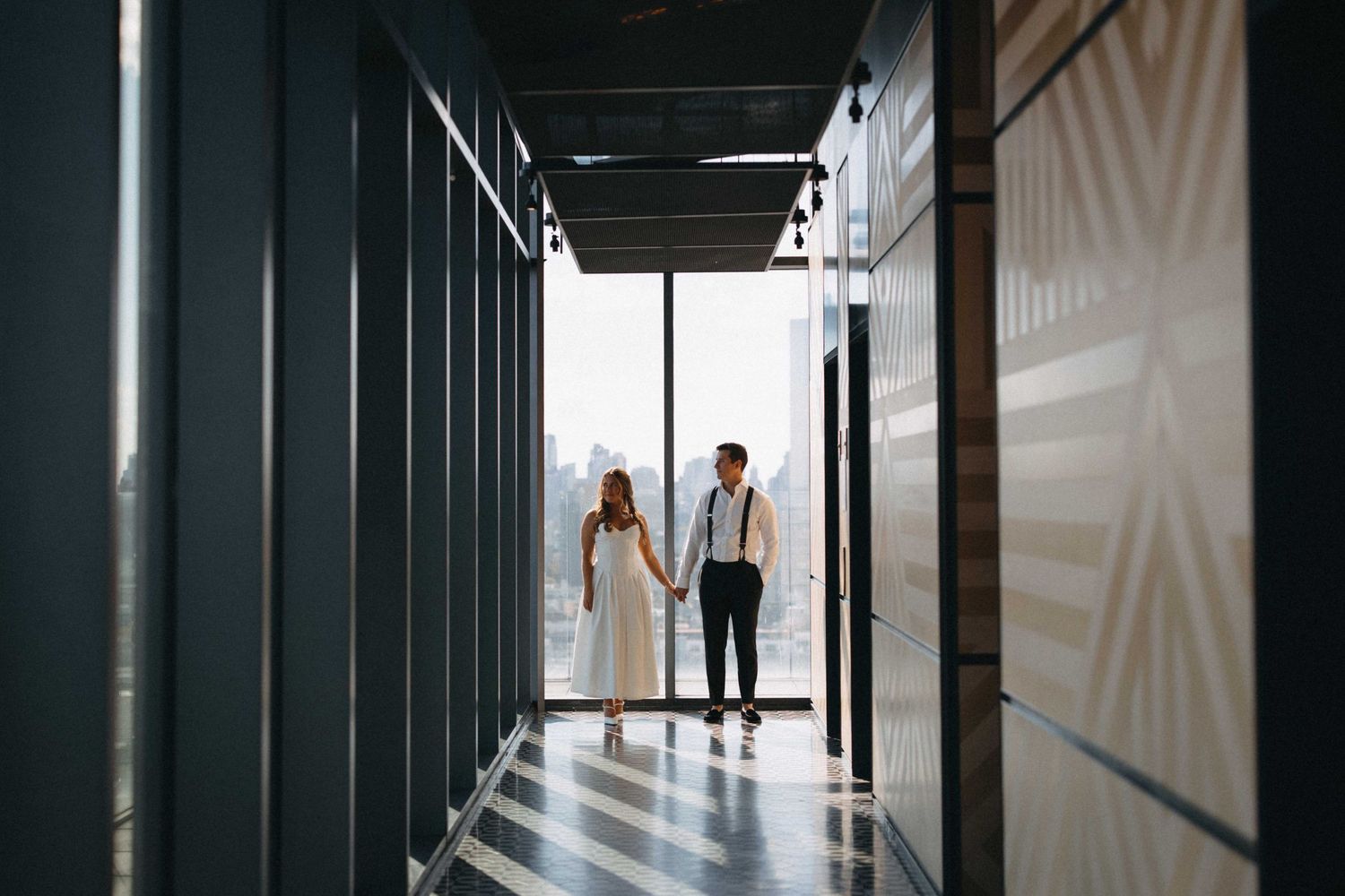 A couple holds hands walking through a sleek modern corridor with a city skyline in the background.