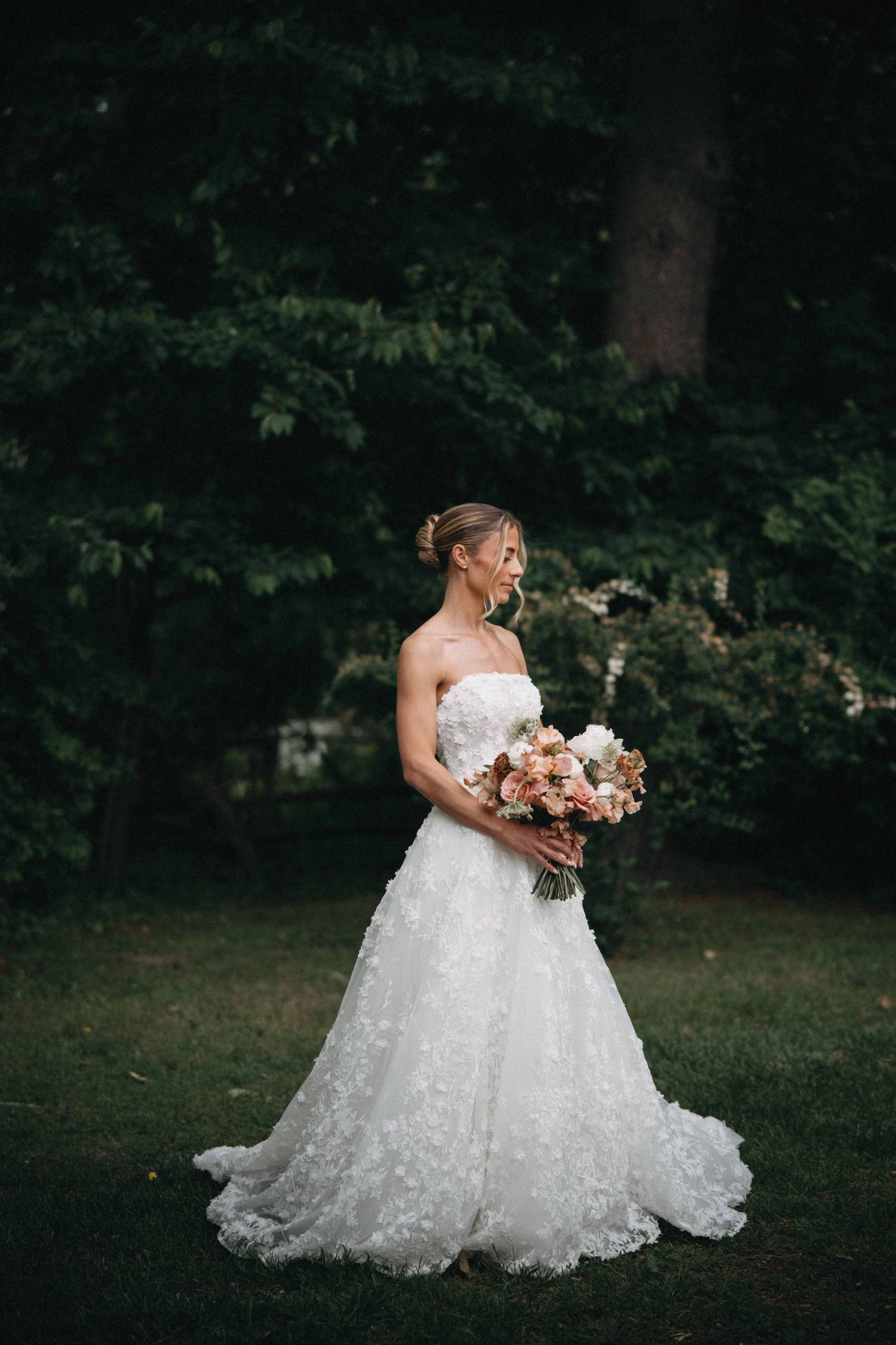 Bride in a strapless floral ball gown holds a blush bouquet while standing gracefully among dark green trees.