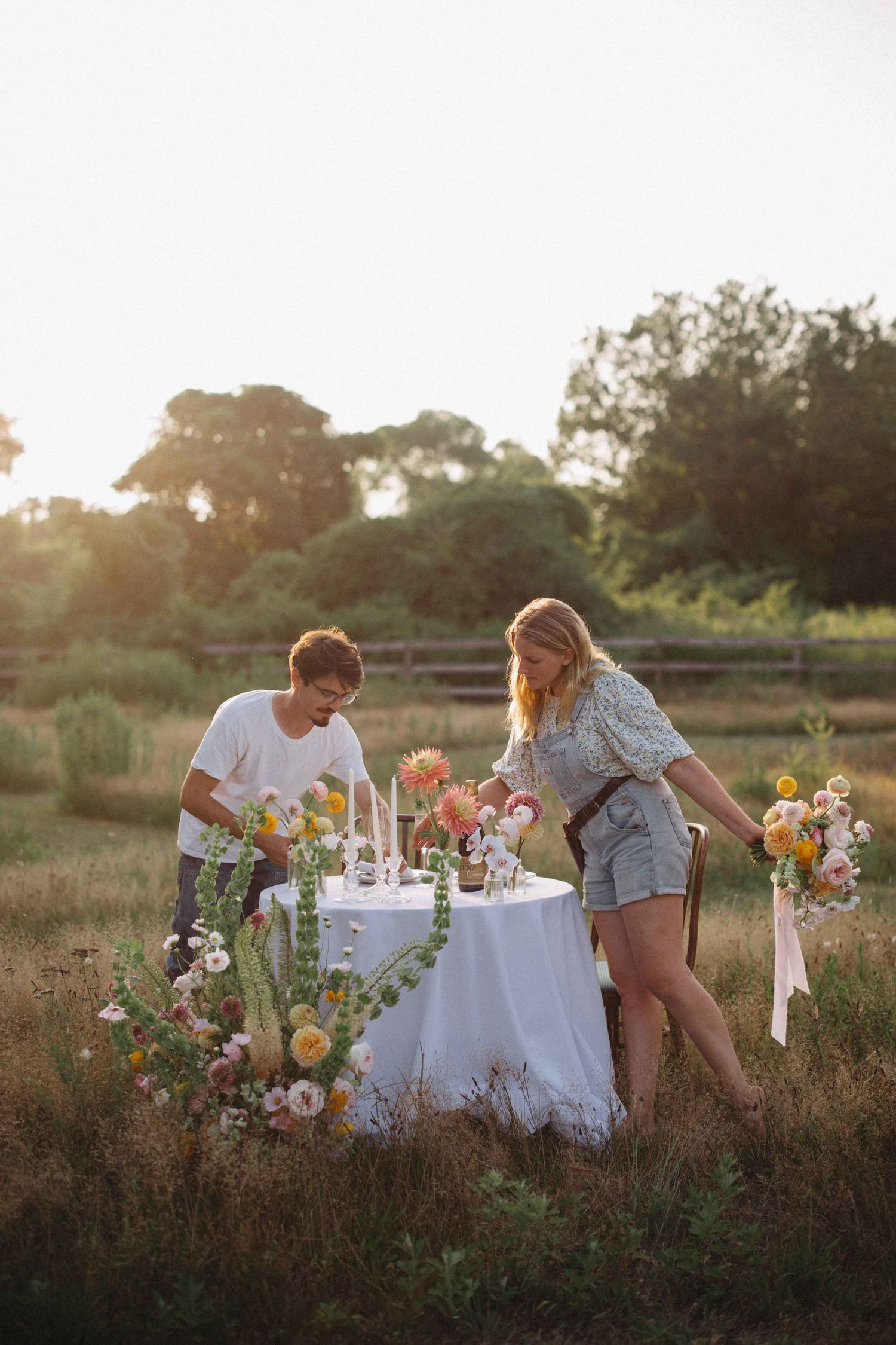 Couple arranging colorful flowers on a white linen table in a sunlit meadow at golden hour.