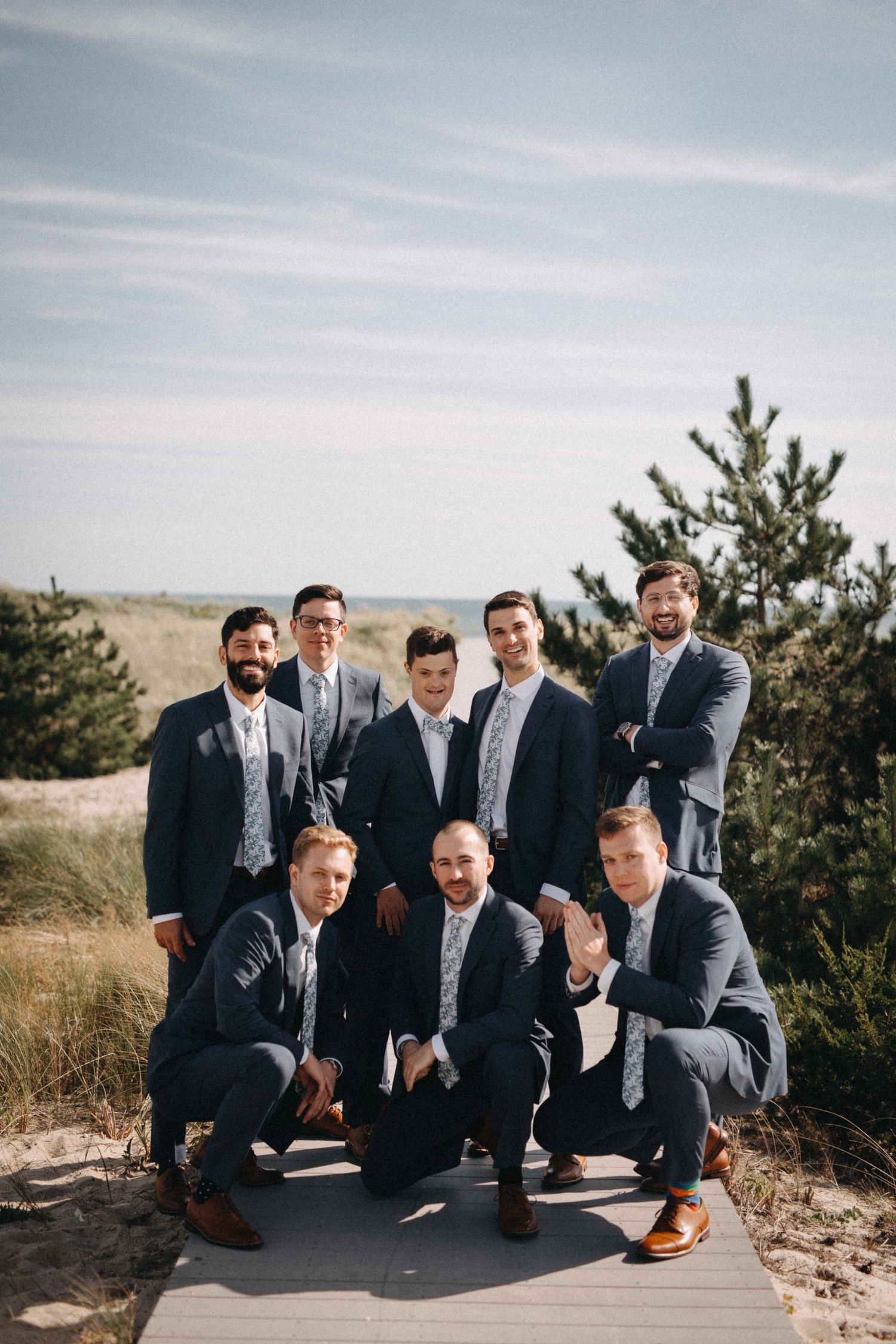 Eight groomsmen in navy suits pose together on wooden boardwalk surrounded by coastal dunes.