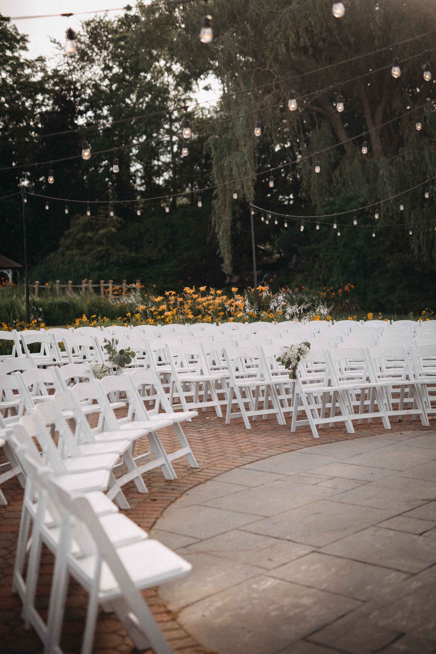 Outdoor wedding ceremony setup with curved rows of white folding chairs, string lights, and yellow flower garden backdrop.