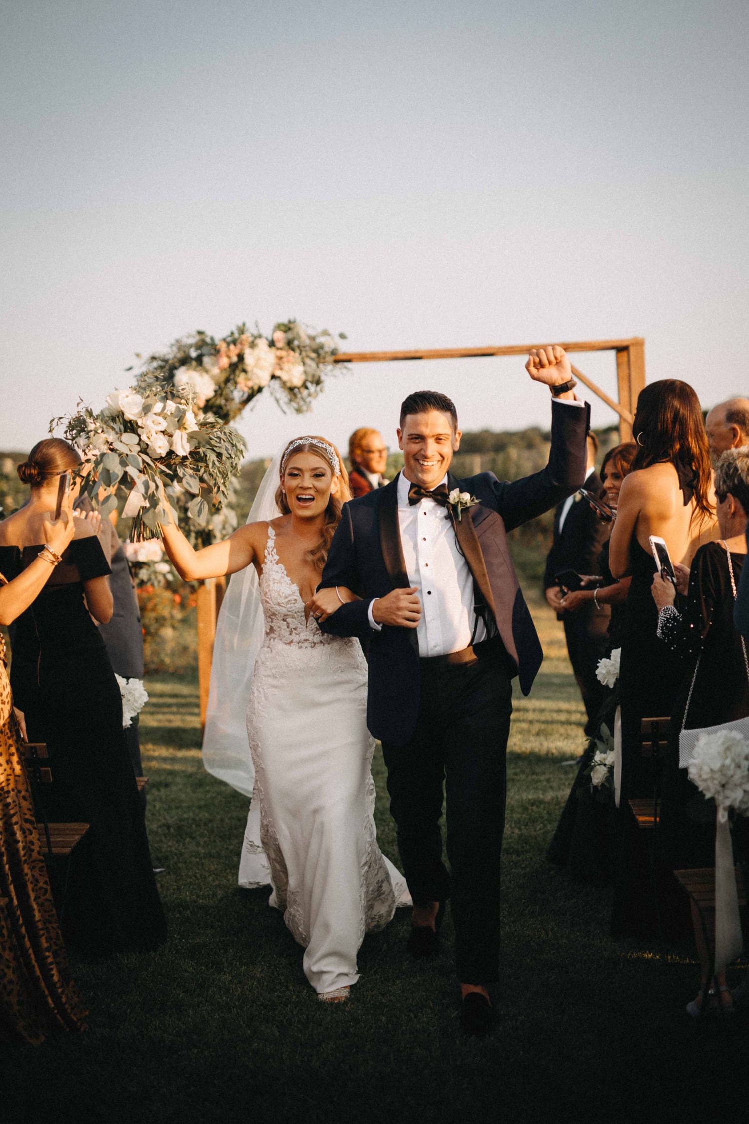 Joyful newlyweds celebrating recessional at outdoor vineyard ceremony with wooden arch backdrop.