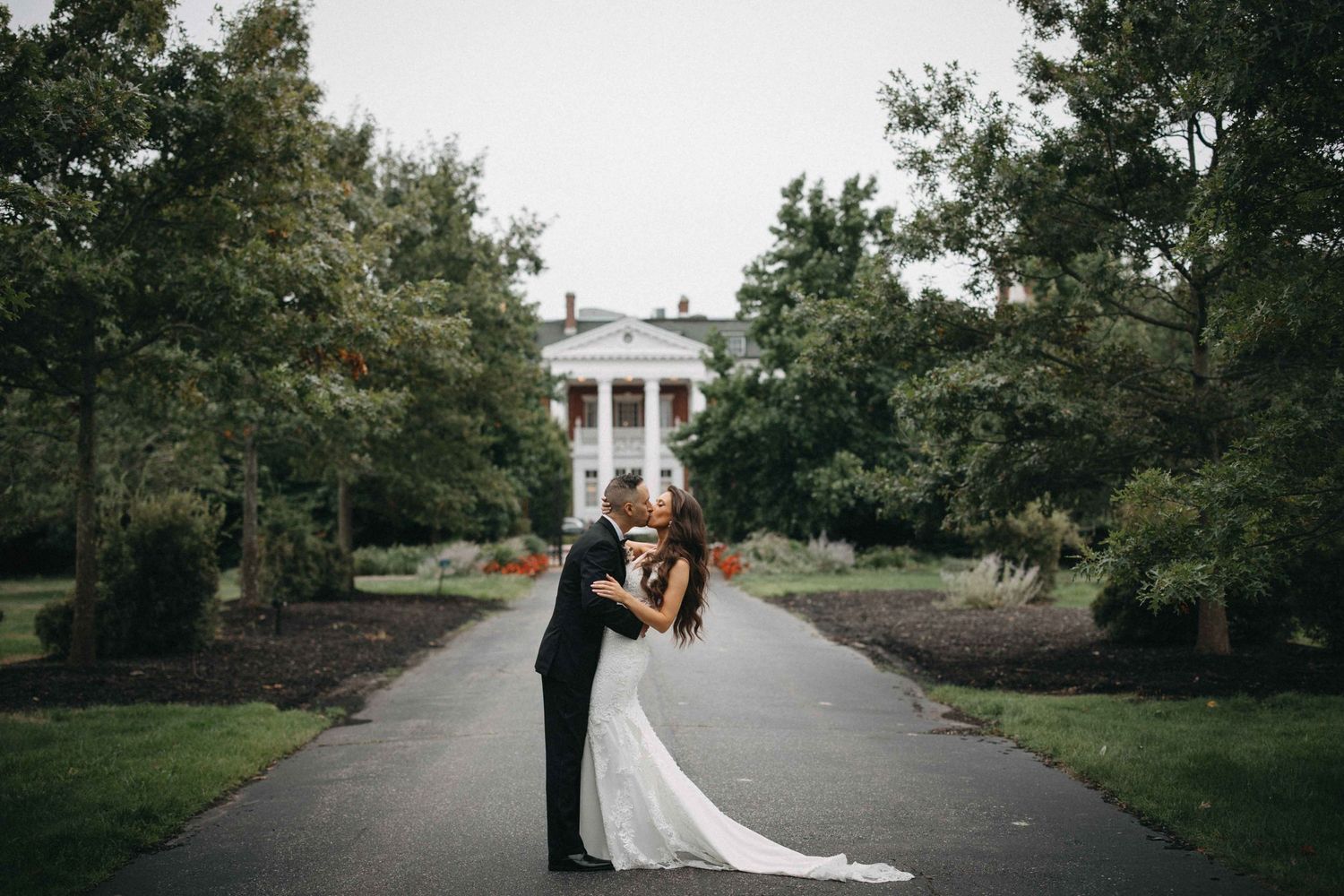 Bride and groom share a romantic kiss on a pathway leading to an elegant white manor.