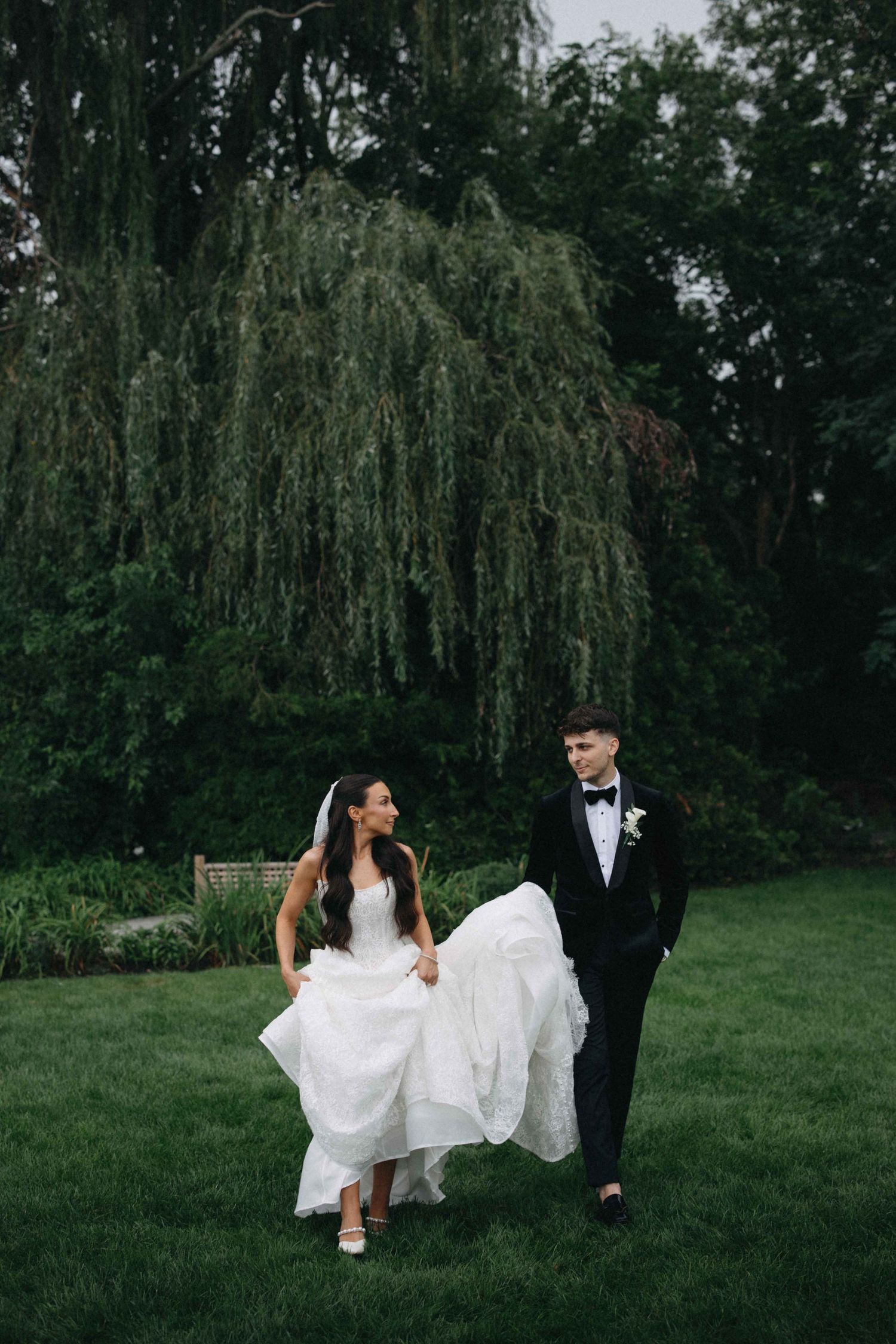 Bride and groom walk hand in hand on lush green lawn before a weeping willow tree during outdoor wedding.
