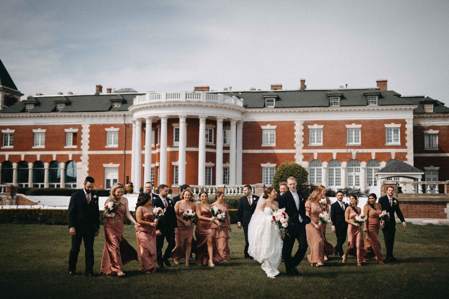 Wedding party poses on lawn before grand red brick colonial mansion with white columns.
