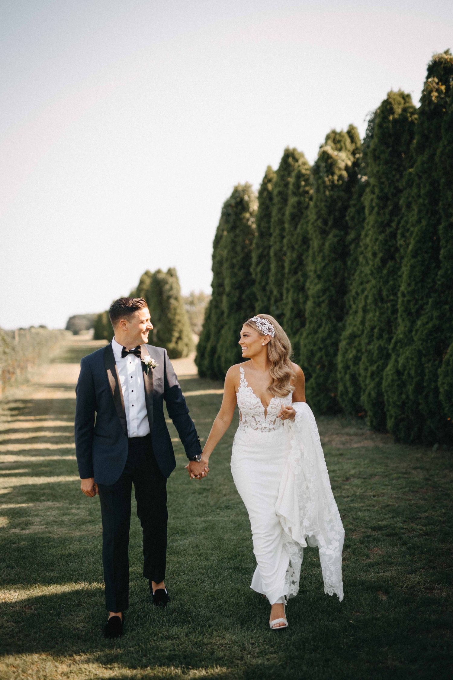 Bride in lace gown and groom in navy tuxedo walking hand in hand by cypress trees.