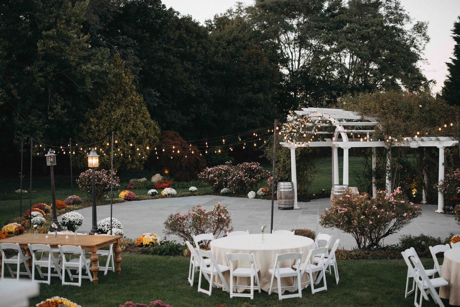 Outdoor wedding reception setup at dusk with round tables, white chairs, string lights, and colorful floral arrangements.