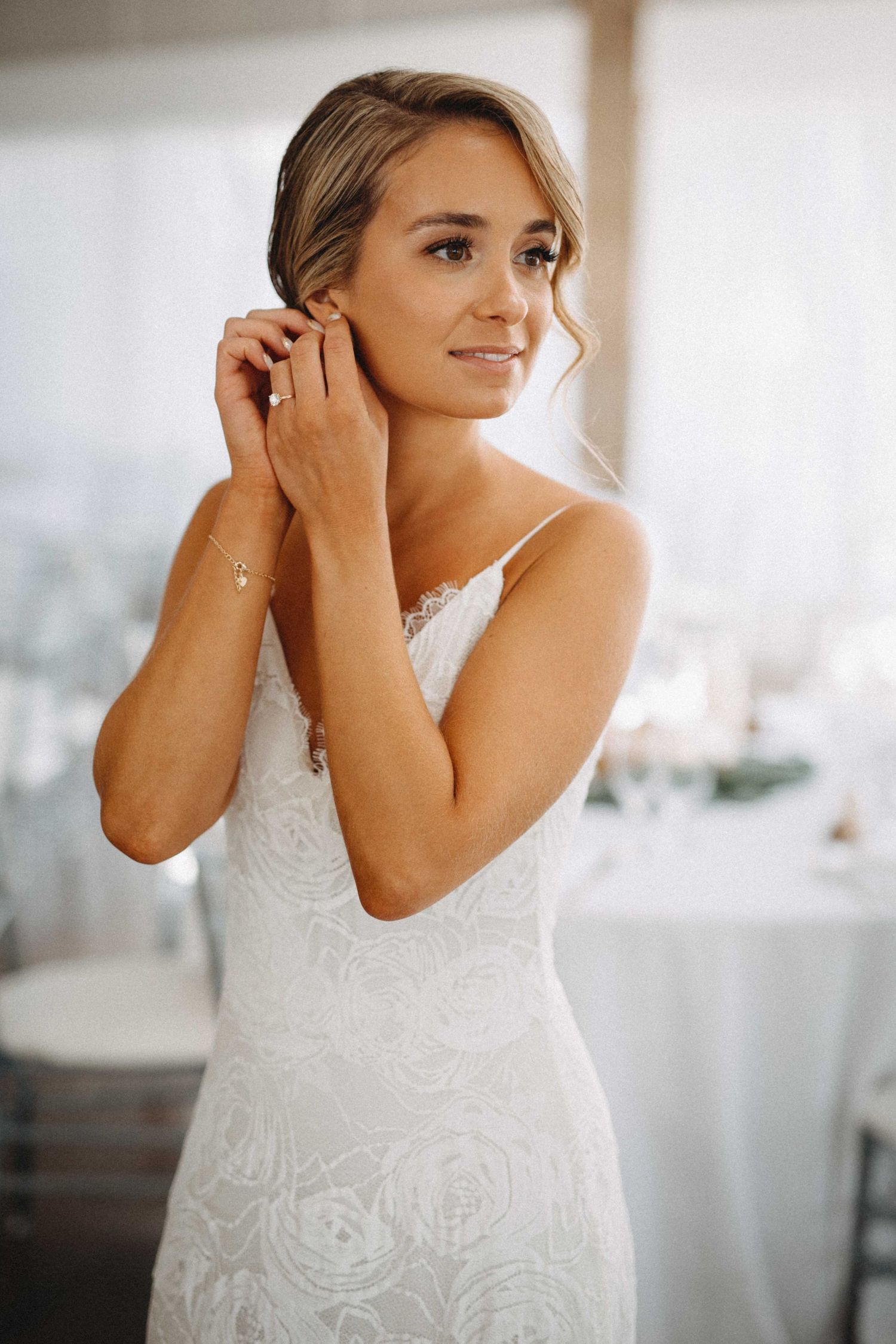 Beautiful bride in lace gown putting on earrings while getting ready for her wedding.
