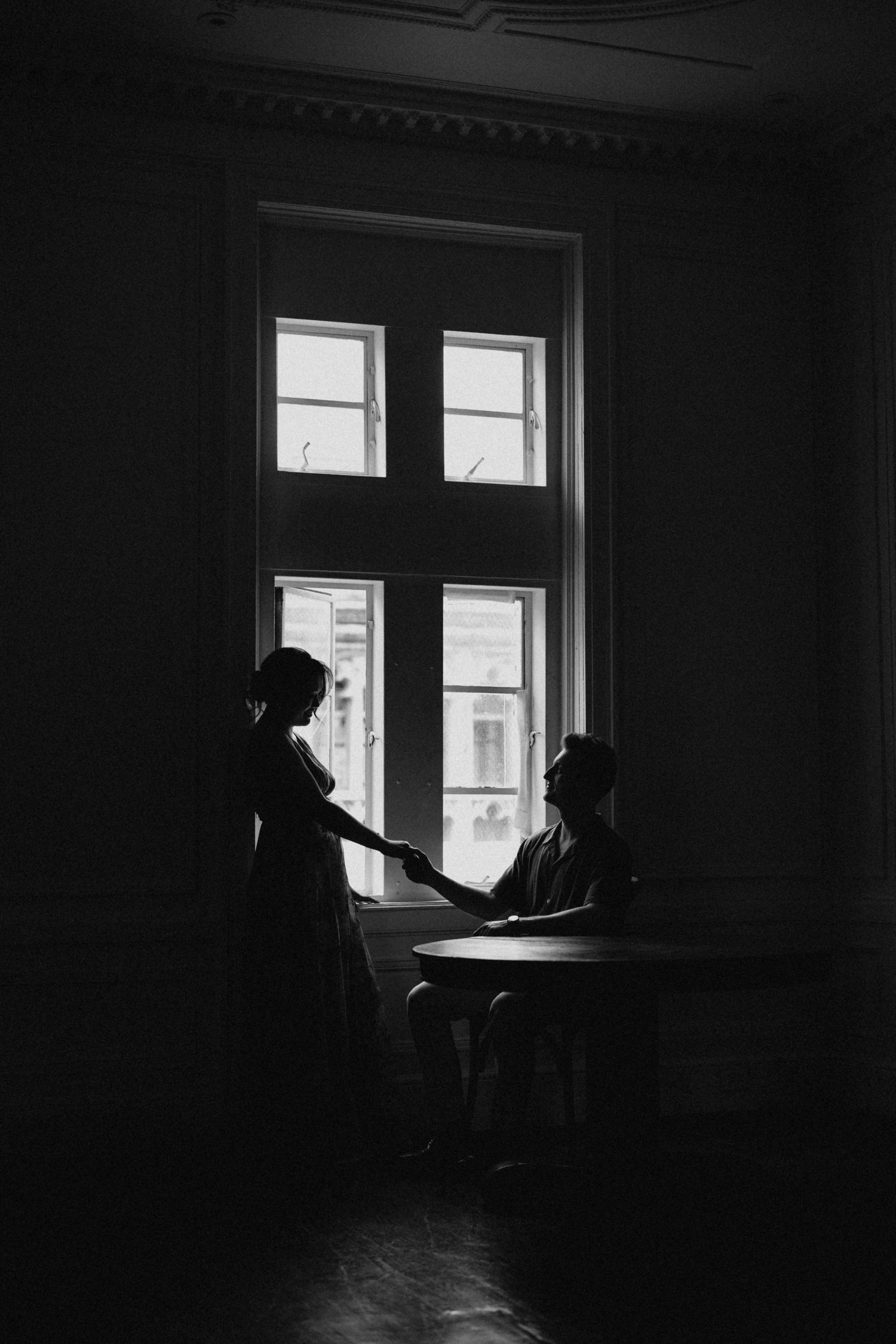 Dramatic black and white silhouette of a couple holding hands by a large window in a dark room.