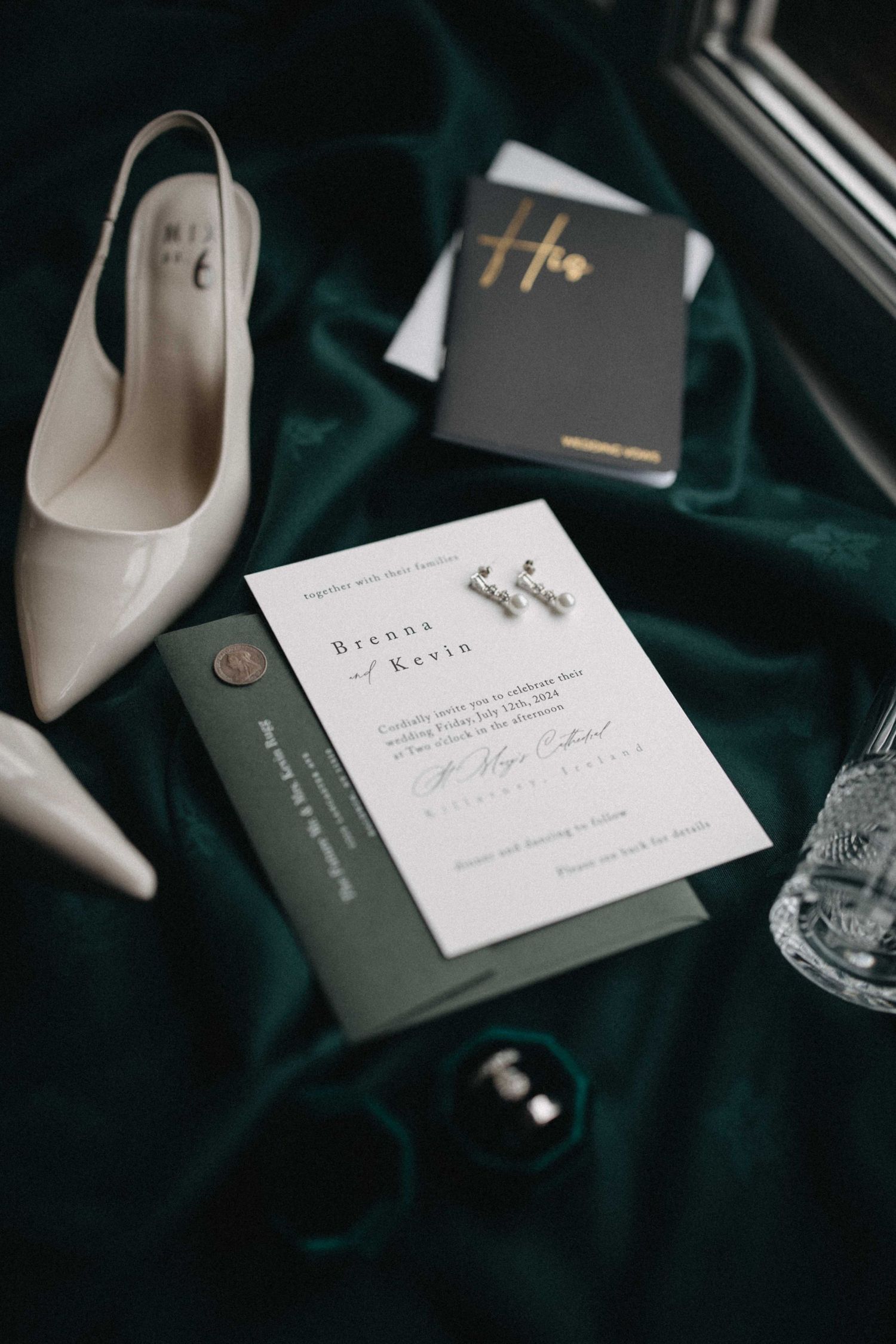 Wedding flat lay featuring an invitation for Brenna and Kevin, white heels, earrings, and rings on green velvet.