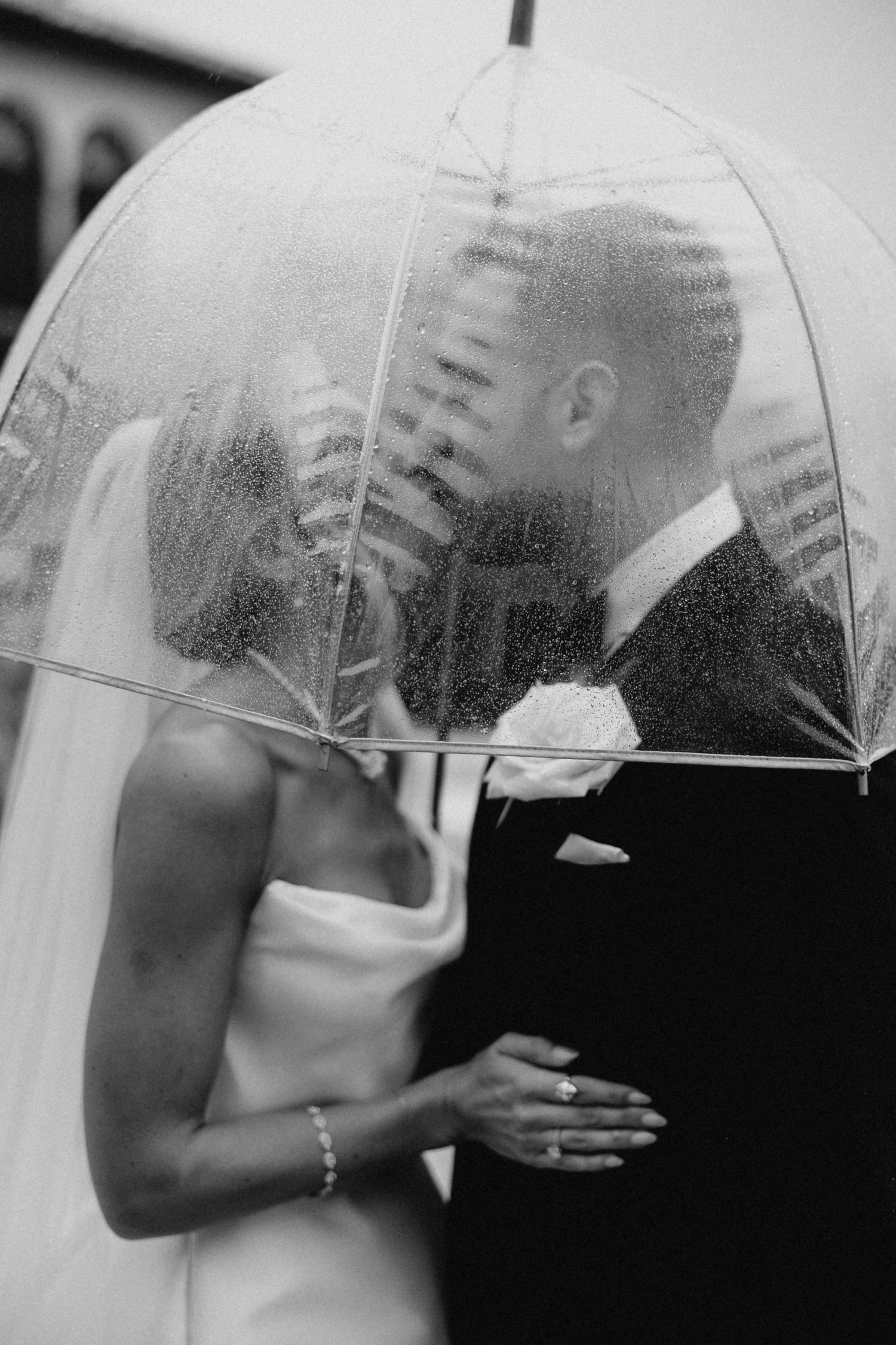 Black and white romantic wedding portrait of couple kissing under clear umbrella in the rain.
