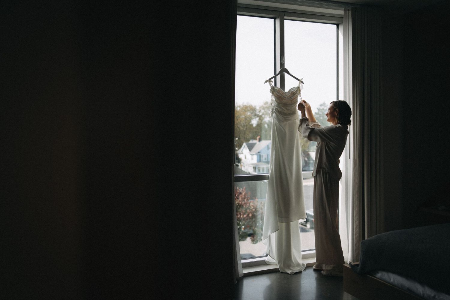 Bride in robe adjusting wedding dress on hanger by bright window in dimly lit bridal suite.