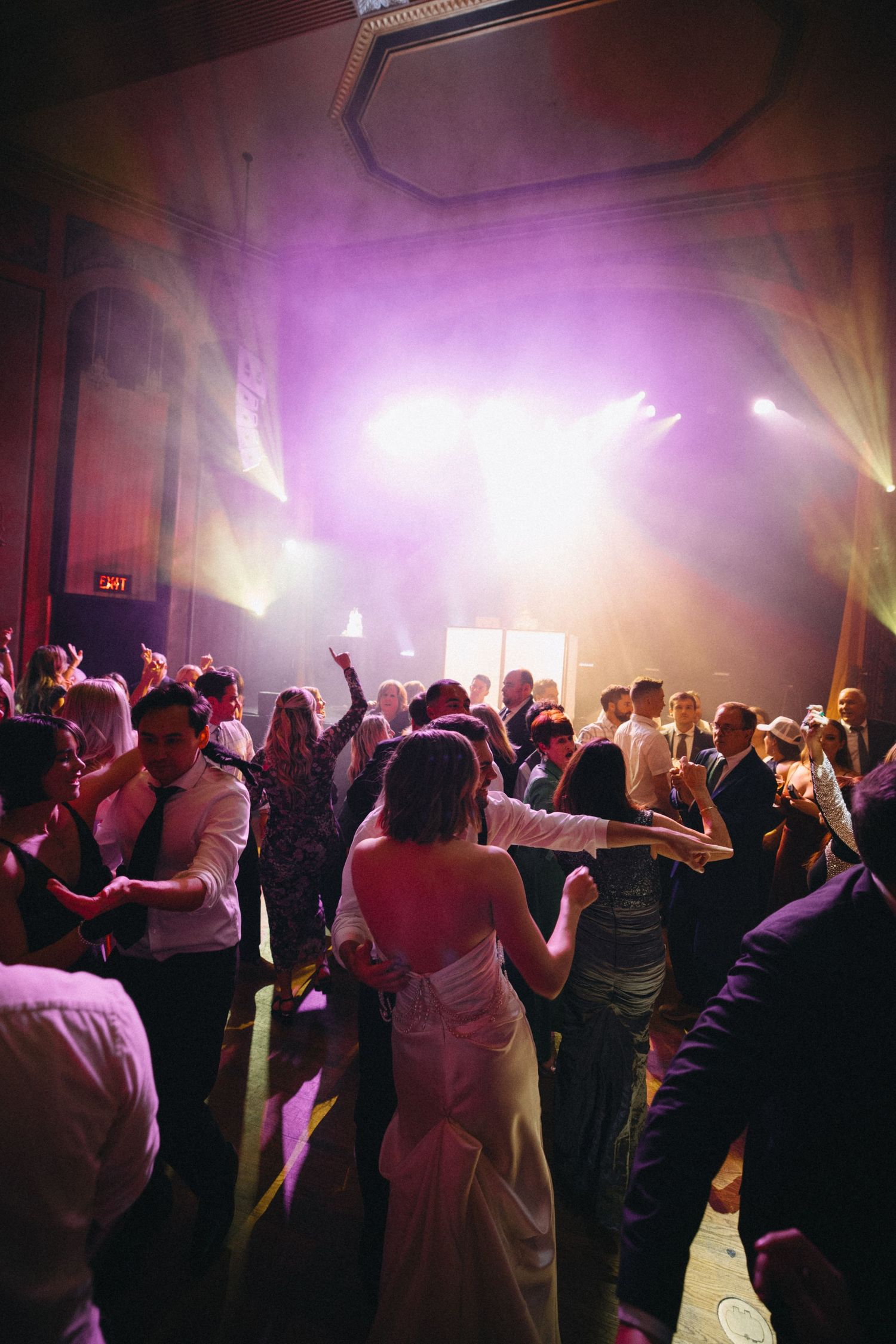 Bride dances joyfully with guests under vibrant purple stage lights at a lively wedding reception.