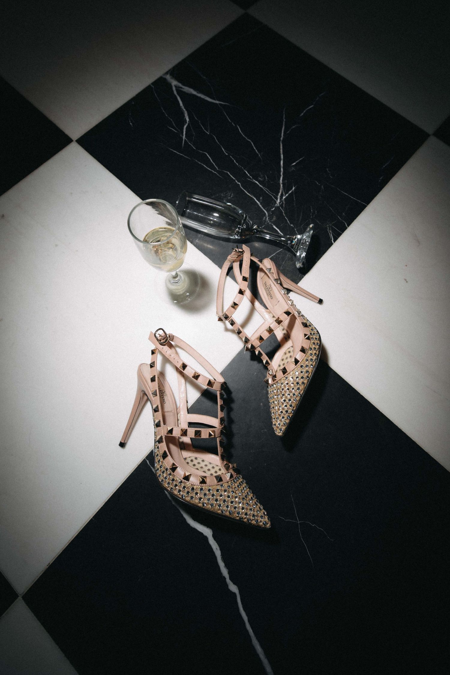 Studded Valentino heels on black and white marble floor beside a glass of champagne.