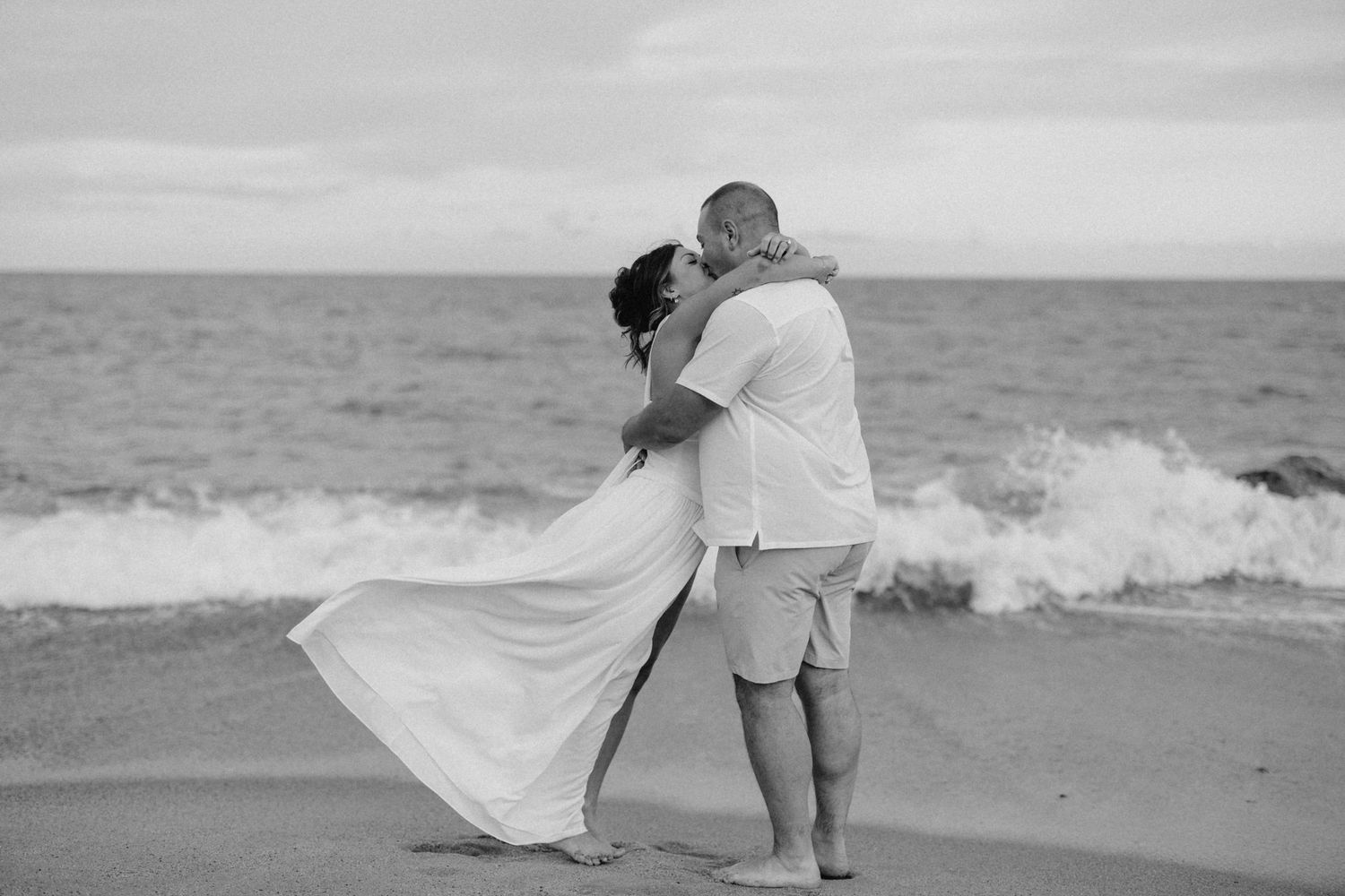 Black and white photo of a couple embracing and kissing on a windy beach shoreline.