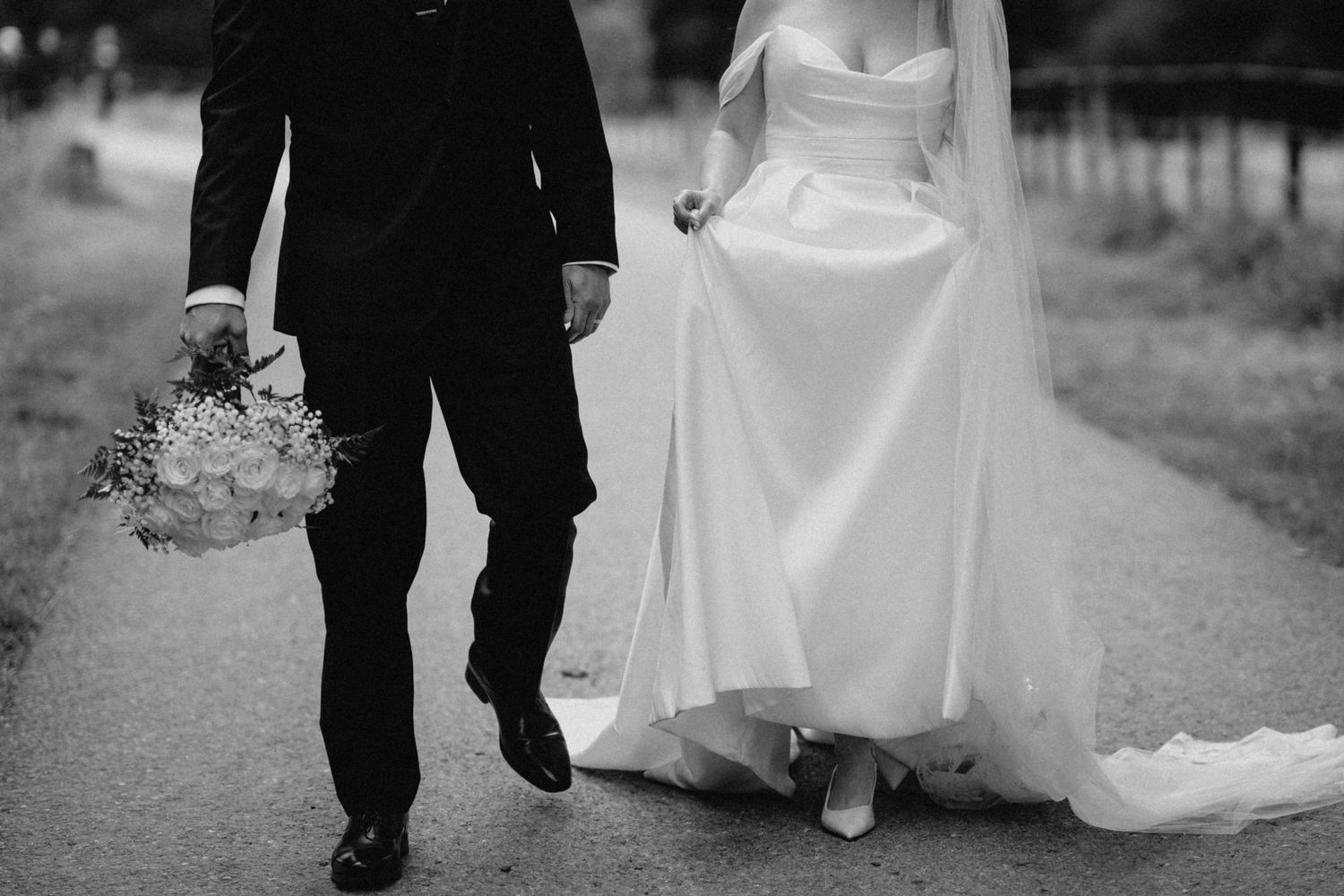 Black and white photo of bride and groom walking hand in hand on garden path.