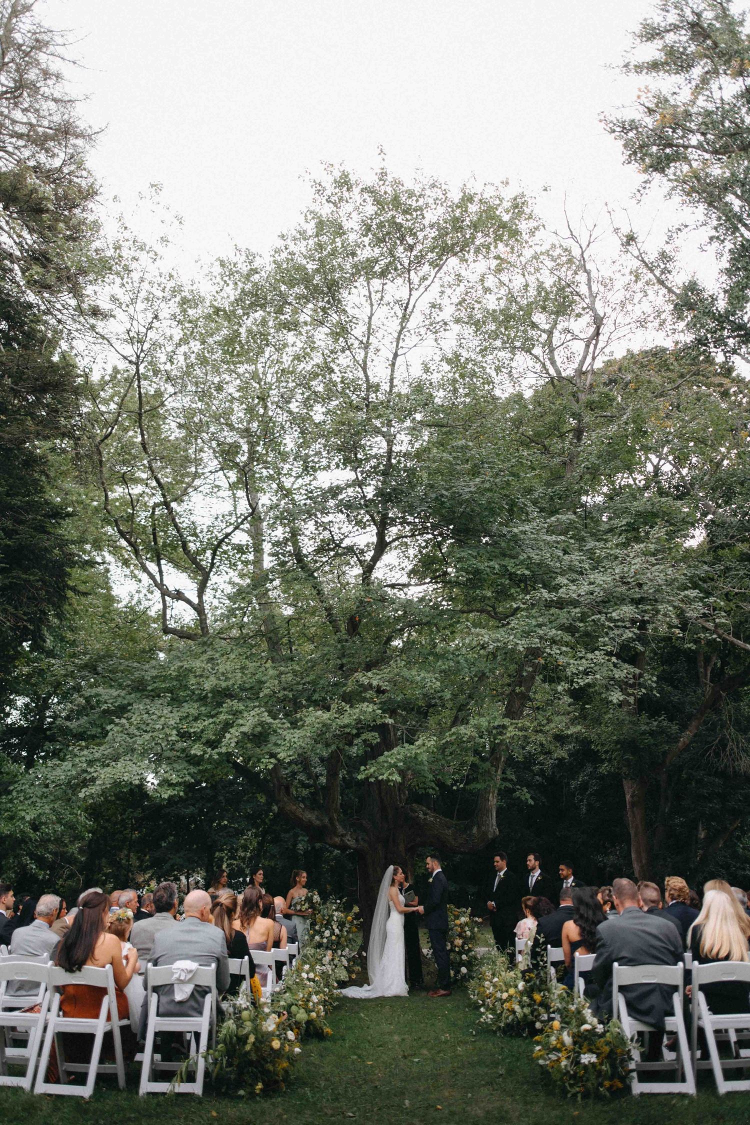 Outdoor wedding ceremony under lush green trees with guests seated in white chairs along a floral-lined aisle.