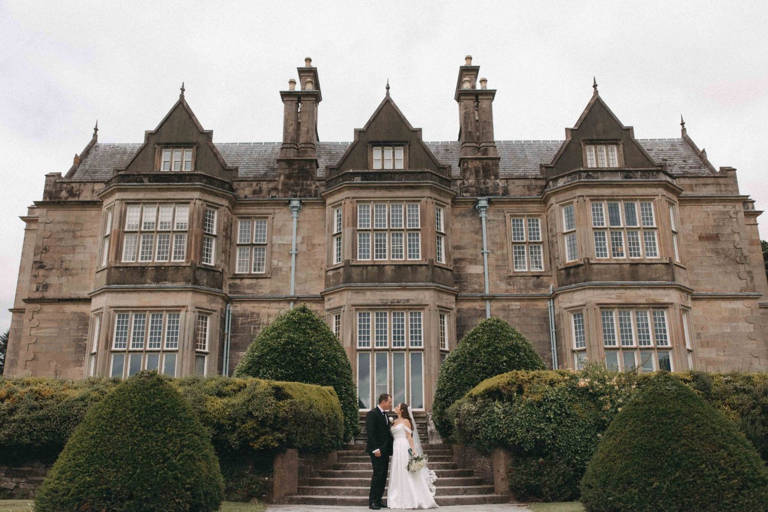Bride and groom pose on steps before a grand Scottish manor house with manicured topiary hedges.