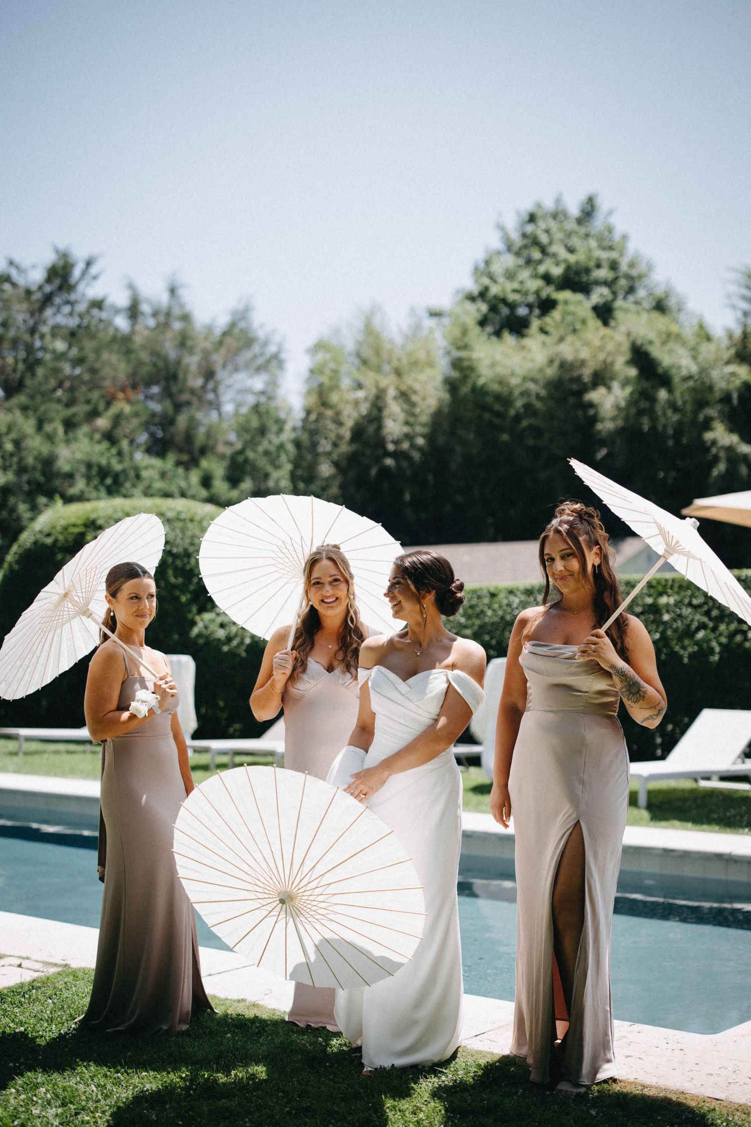 Bride with three bridesmaids in taupe gowns holding white parasols standing poolside on a sunny wedding day.
