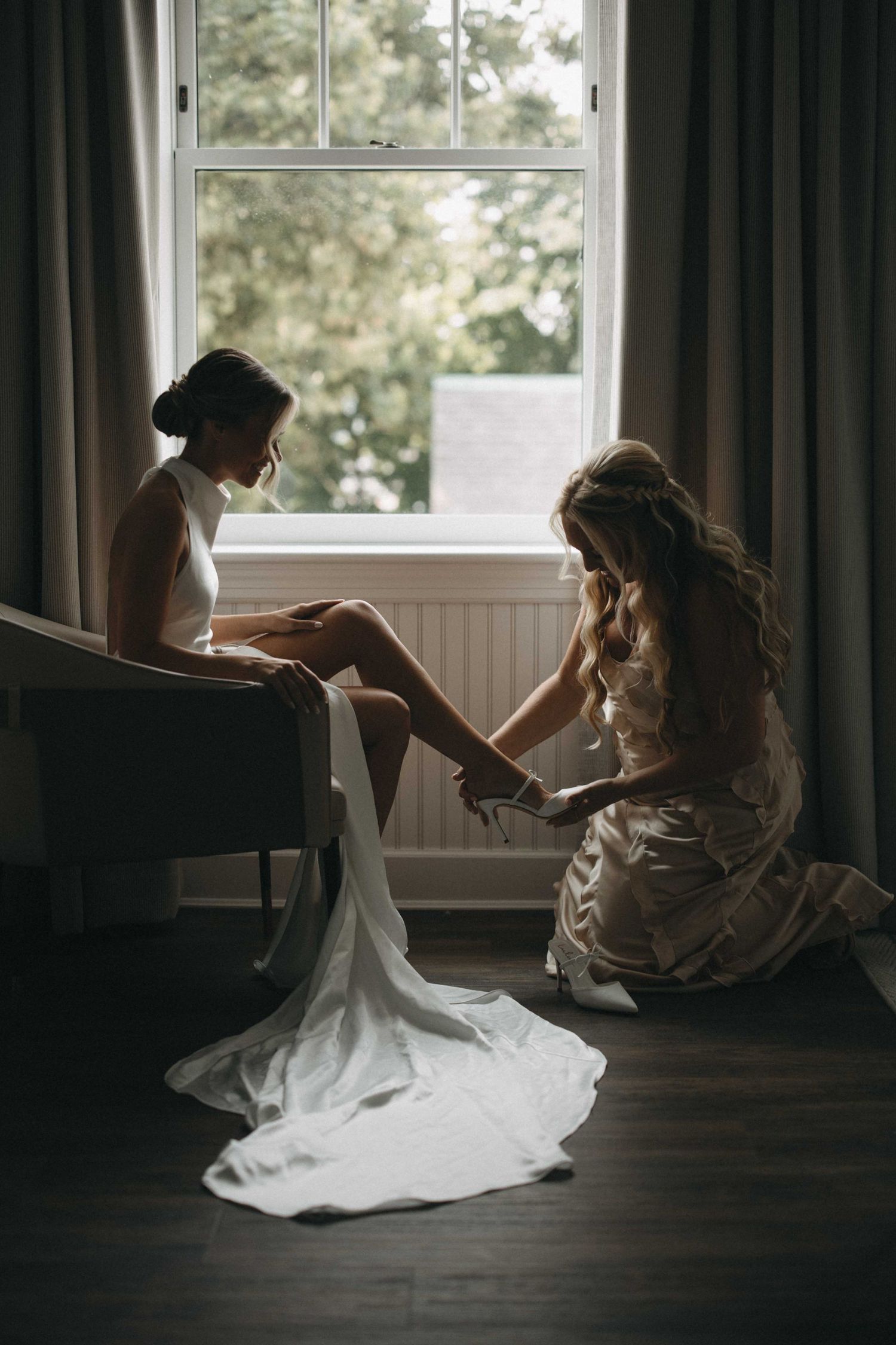 Bridesmaid kneels to help bride put on wedding shoes by a bright window before the ceremony.