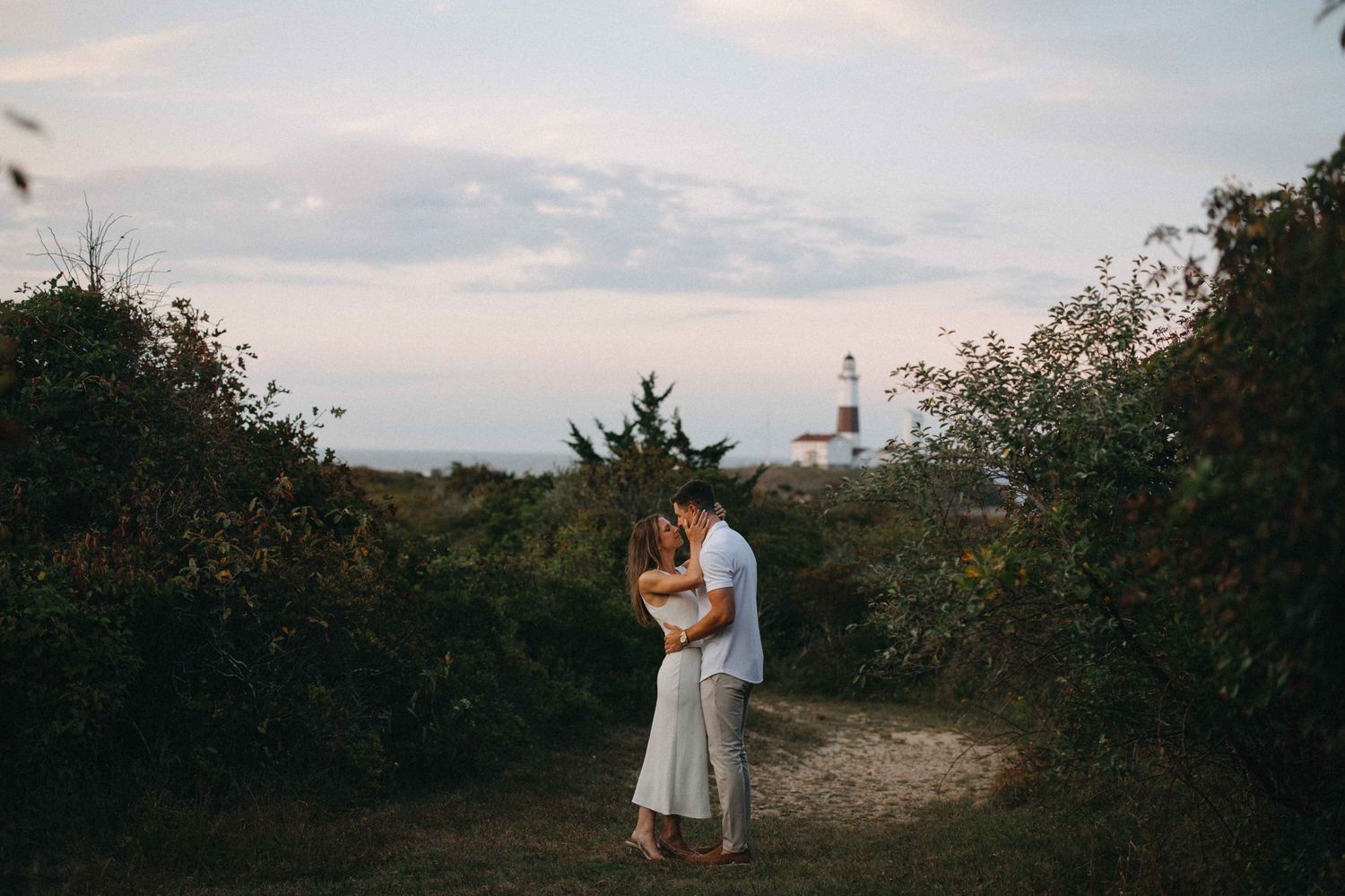 Couple embracing on a nature path with a lighthouse visible in the background.